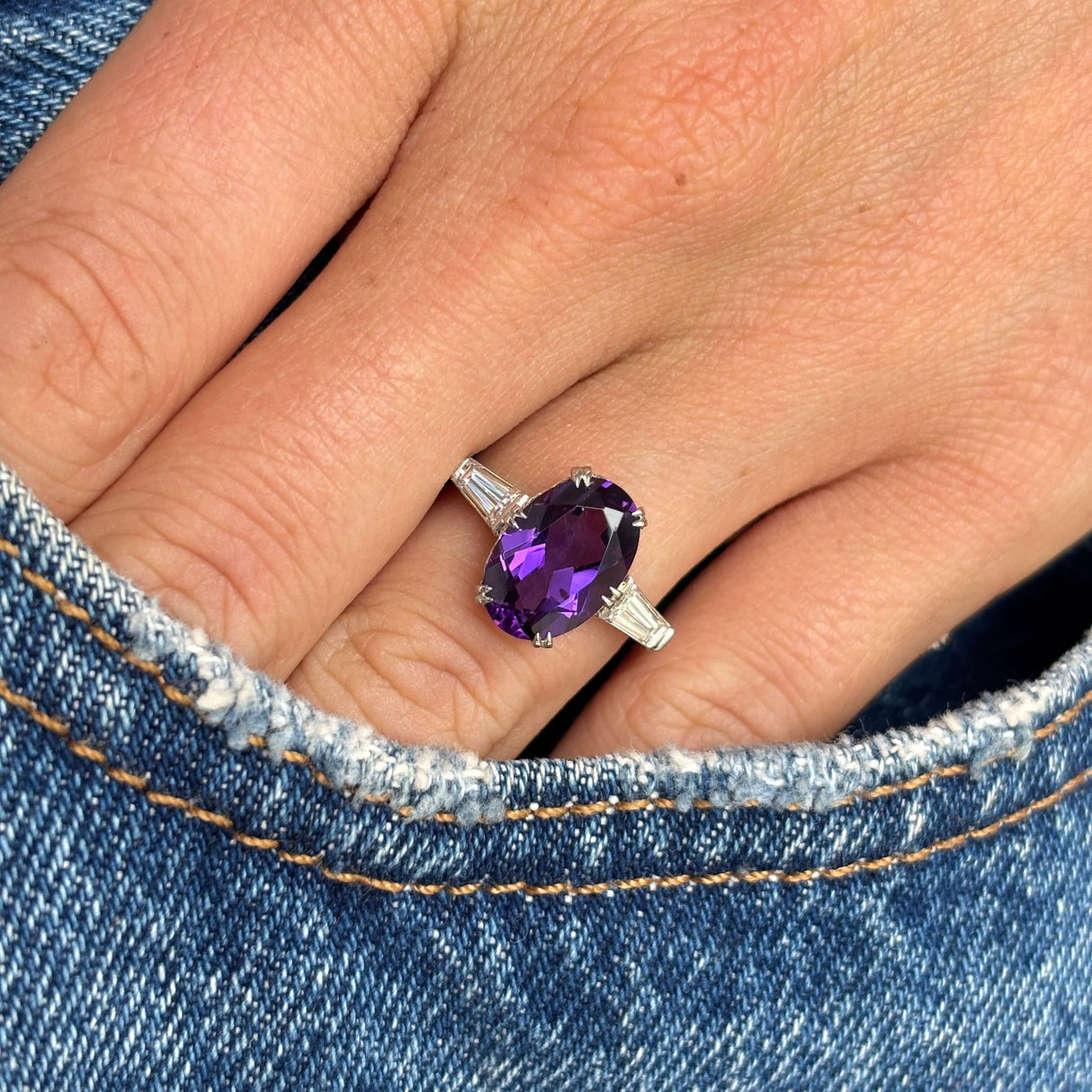 Antique Art Deco amethyst and diamond three-stone ring in platinum styled on hand against blue denim, showcasing the richly coloured amethyst centre stone and elegant vintage design.