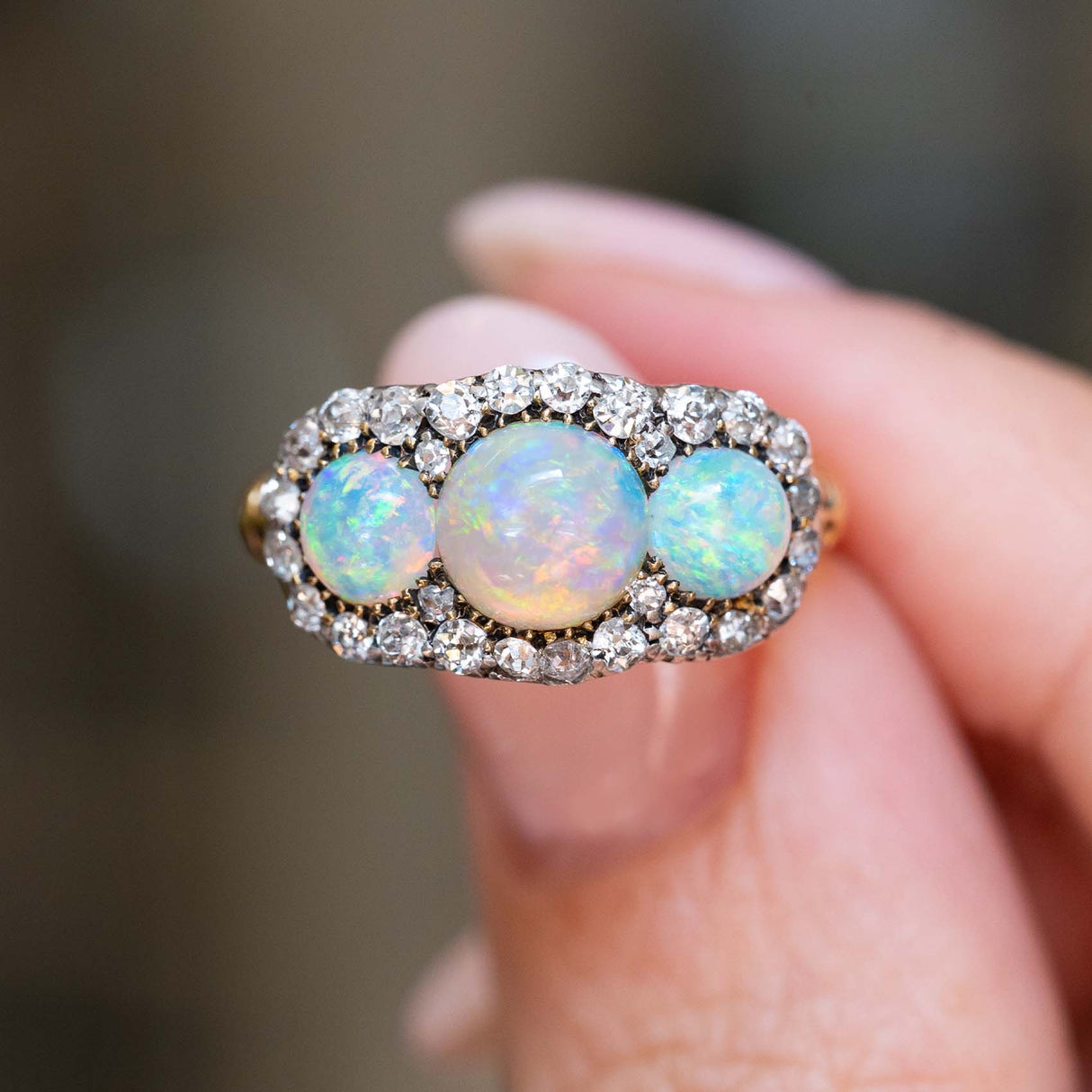A close-up of an antique three-stone opal & diamond cluster ring shown between fingers.