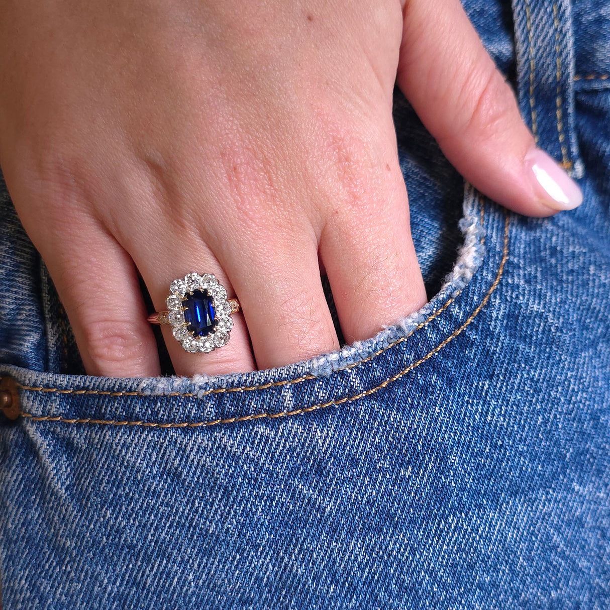 Detail of a hand wearing sapphire and diamond cluster ring tucked into a pocket of denim jeans