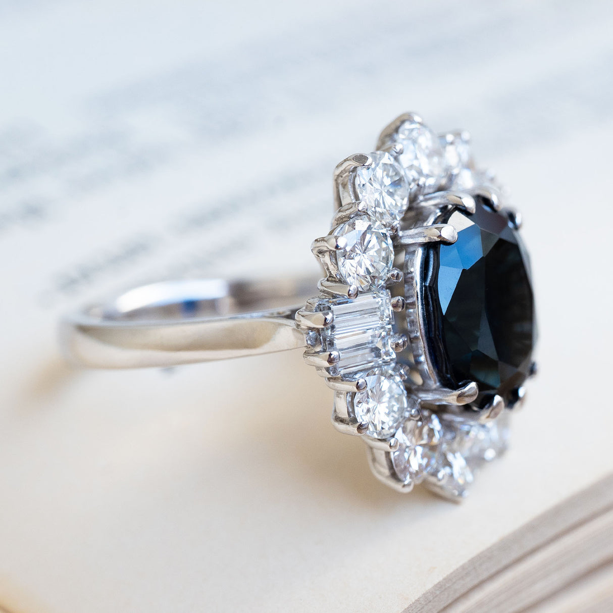 A close-up of a platinum vintage ring with an oval blue sapphire in the centre flanked by two baguette-cut diamonds.