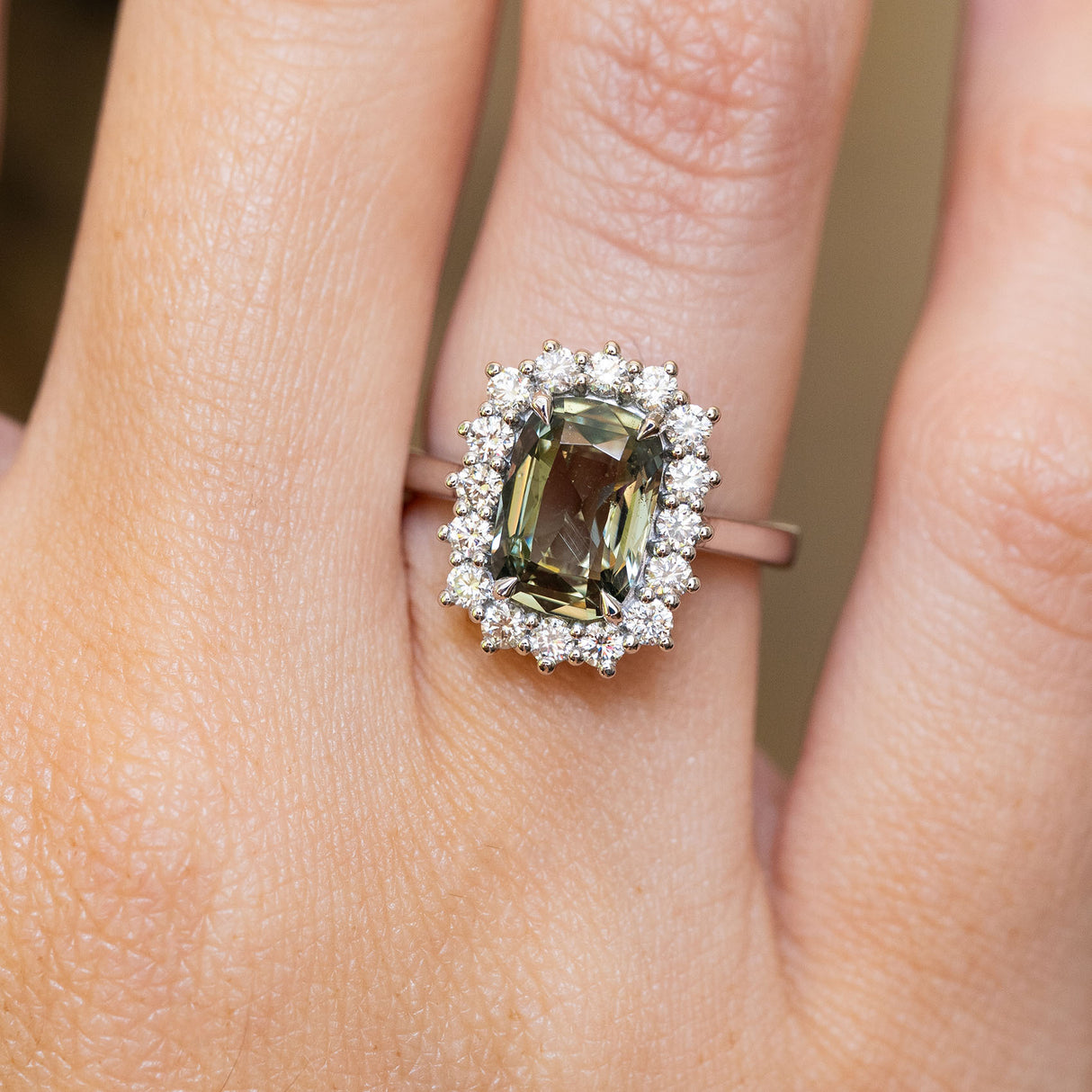 A close-up of a hand wearing an antique-inspired, green cushion-cut sapphire and diamond halo engagement ring.