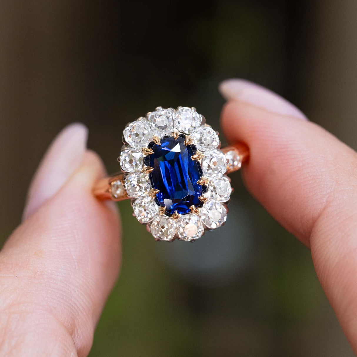 An antique Jugendstil royal-blue sapphire and diamond cluster ring held between the fingers.