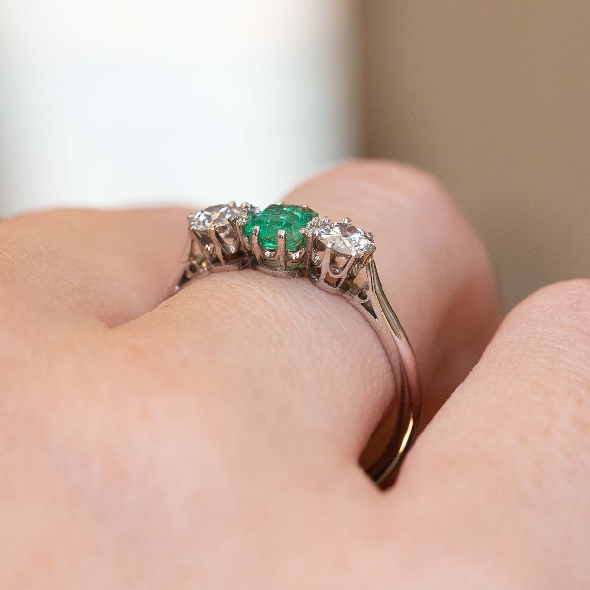 A close-up of a ring worn on a finger, featuring a central green emerald gemstone flanked by two round diamonds in platinum and white gold.