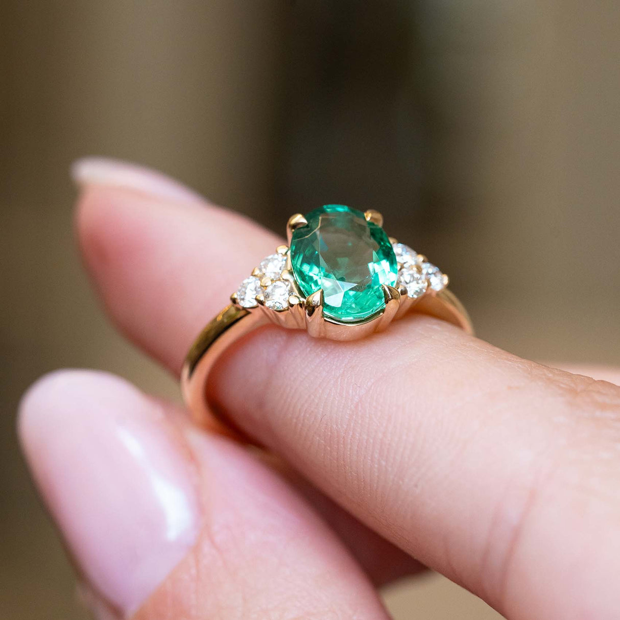 A vintage-inspired oval-cut emerald and diamond trefoil engagement ring held between the fingers.