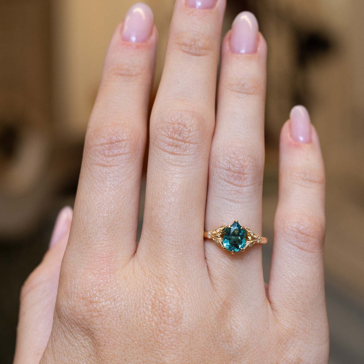 A hand wearing a gold ring with a pear-cut teal tourmaline, set against a softly blurred warm brown background.