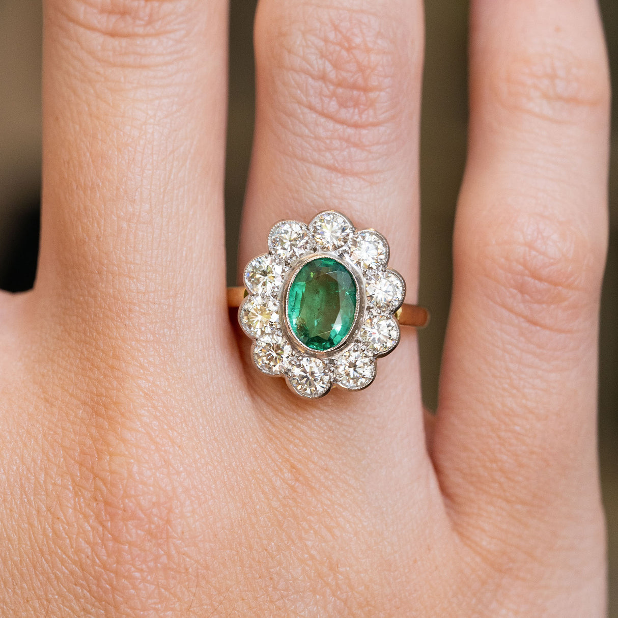 A close-up of a hand wearing a vintage yellow and white gold ring with a green emerald gemstone and diamond cluster ring.