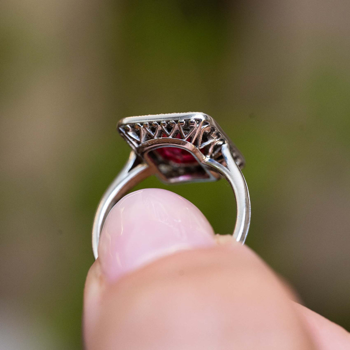 Gallery detail of an antique Art Deco red spinel and diamond cluster ring held between the fingers.