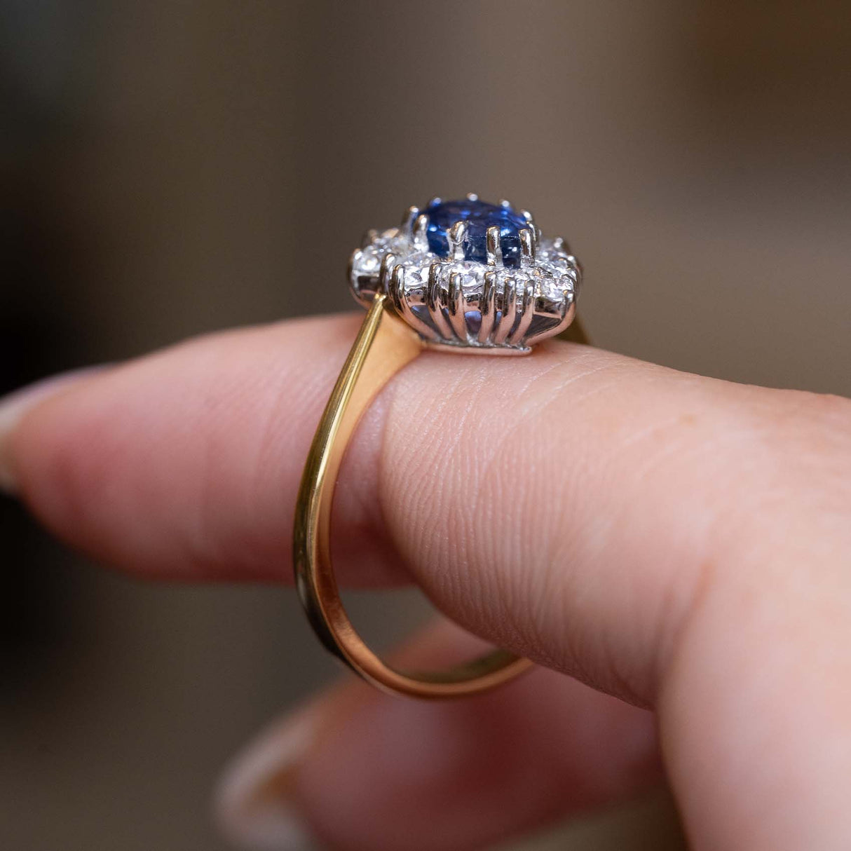 A close-up of a ring being worn on a finger, featuring a blue sapphire center stone surrounded by small diamonds and set on a yellow gold band.