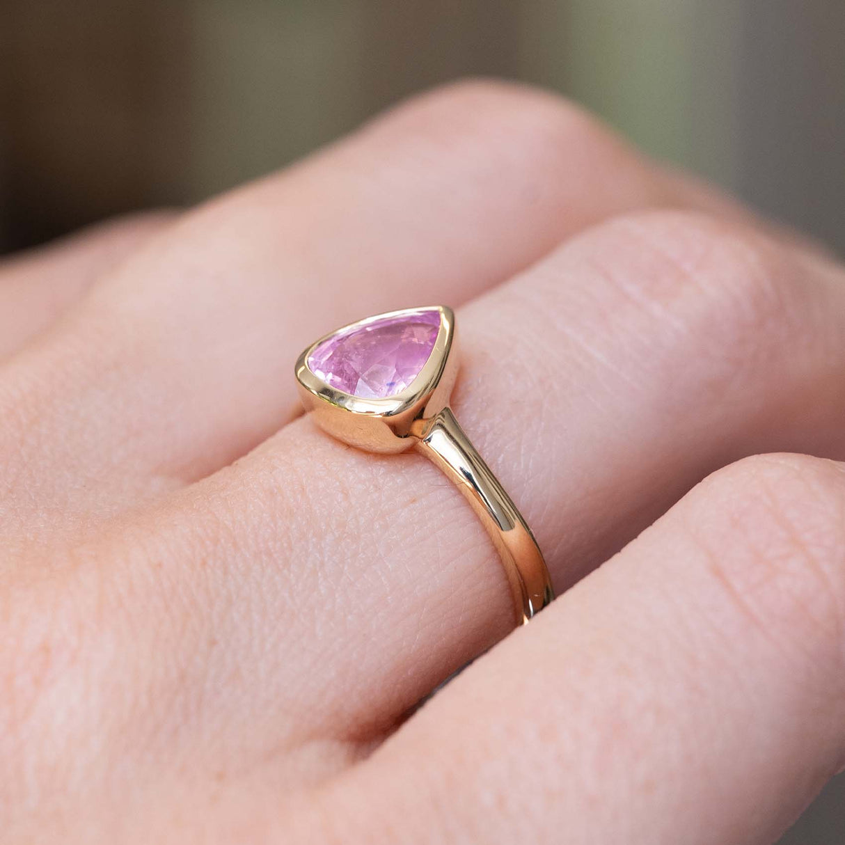 A detailed photo of a yellow gold ring with a drop shaped light pink sapphire drop-cut and rub-over set on an antique inspired solitaire engagement ring, shown from an angle to show how it fits on the finger.