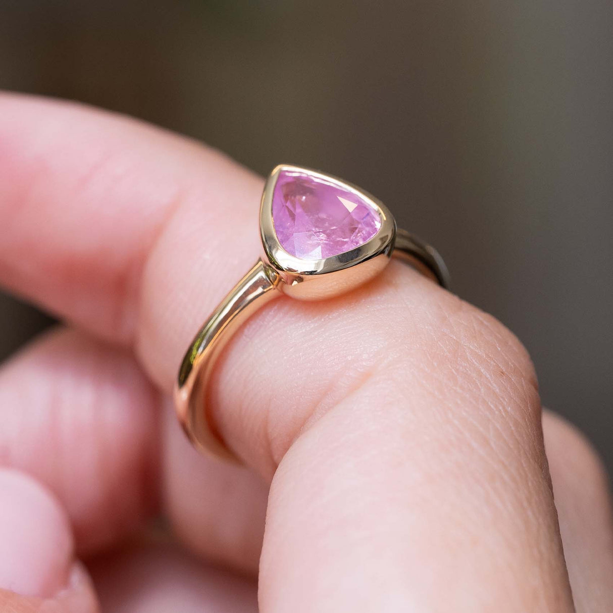 A Antique-inspired, drop-shaped Pink sapphire in a rub-over solitaire engagement ring balanced on a finger shown close up against a blurry background.