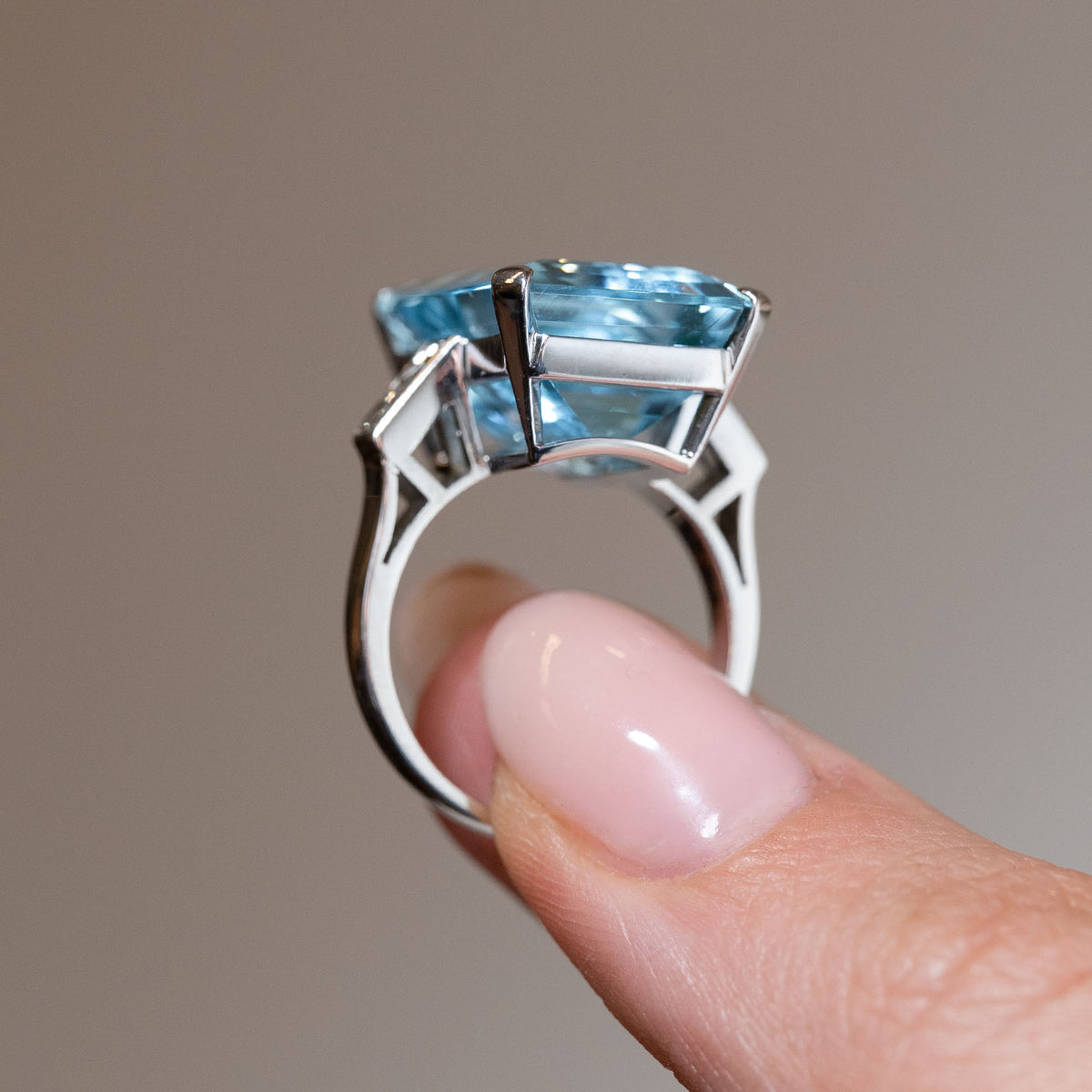 Side profile of a vintage 1970s aquamarine and diamond cocktail ring held between fingers, showing the elevated gemstone, architectural open gallery and geometric white gold shoulders.