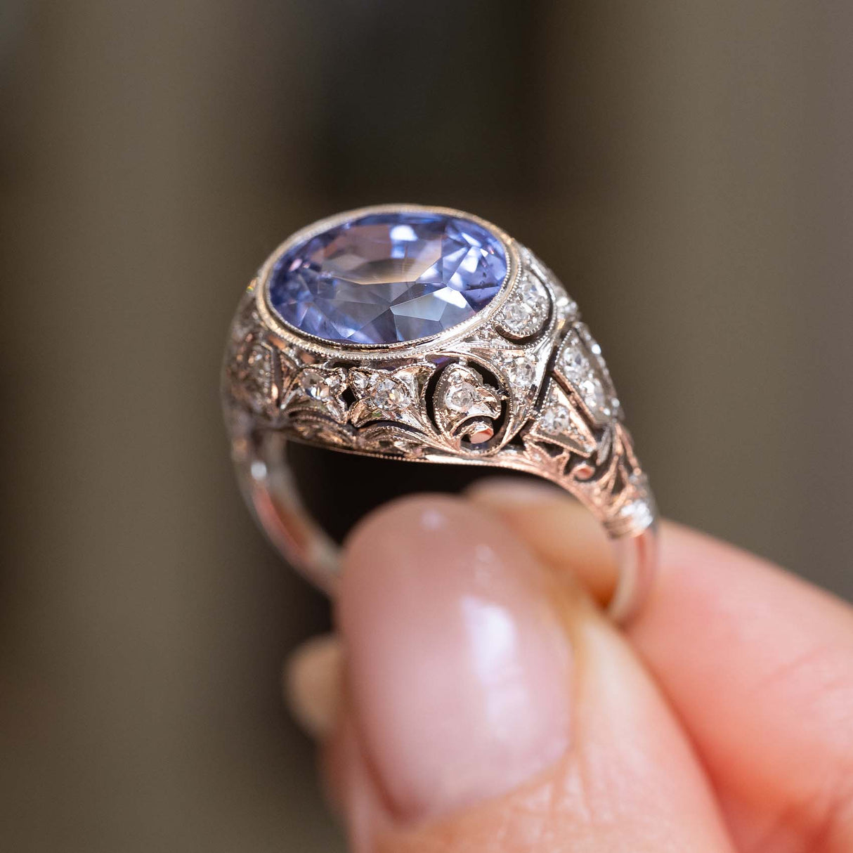 a detailed close image of the ornate details of an Antique, Belle Époque Ceylon colour-change sapphire and diamond cluster ring in platinum against a blurred background.
