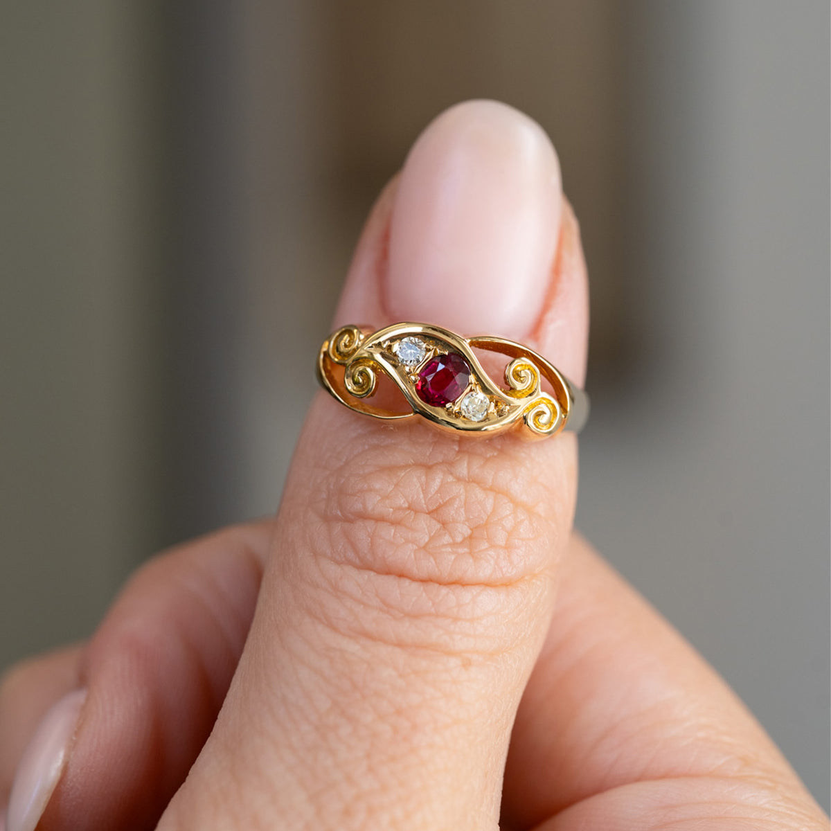 Handheld portrait of the ruby and diamond three-stone ring, displaying the decorative openwork and curled gold details.