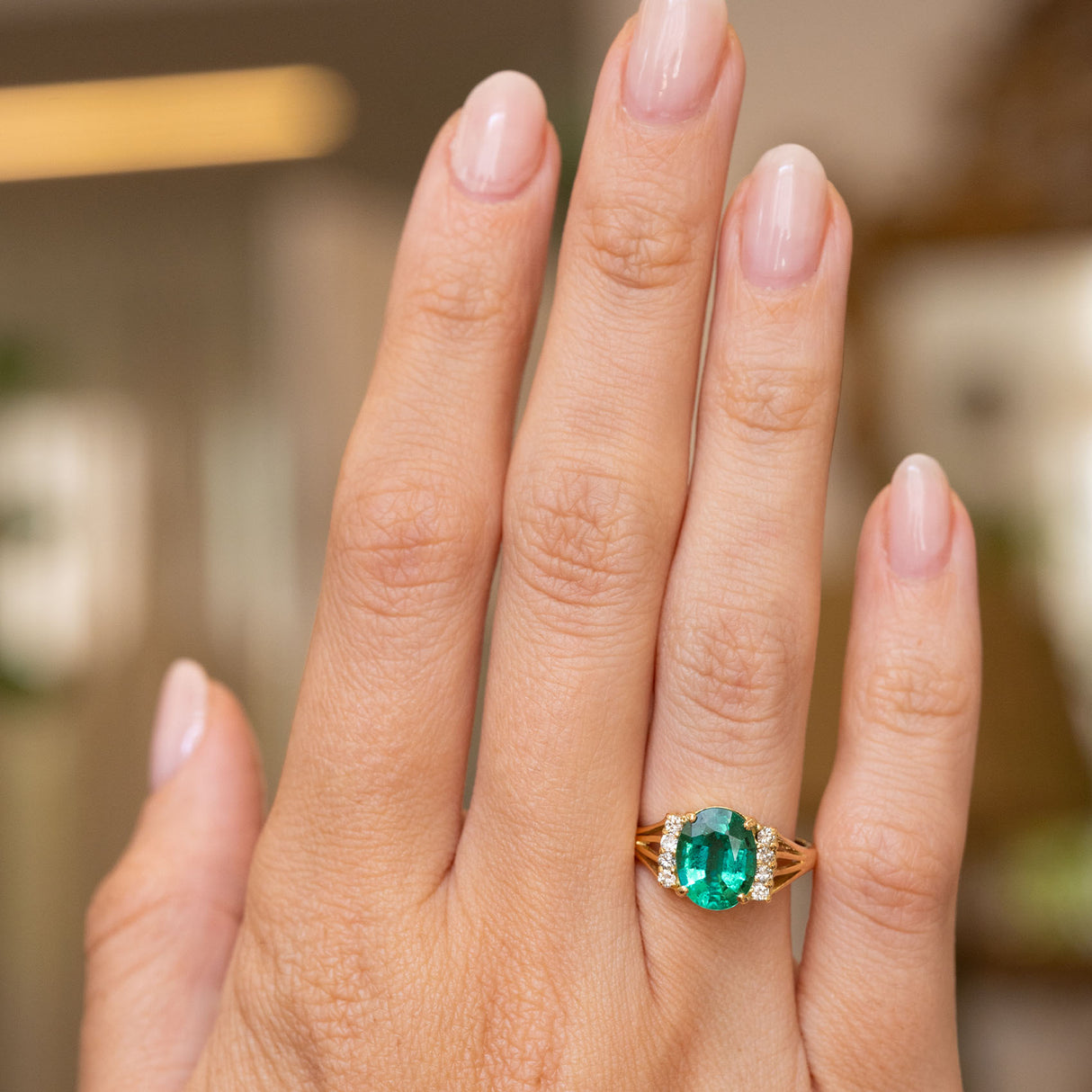 A close-up of a hand wearing a Vintage 1970s yellow gold engagement ring with an oval-cut green emerald centre stone and diamond accents on the shoulders, set against a softly blurred indoor background.