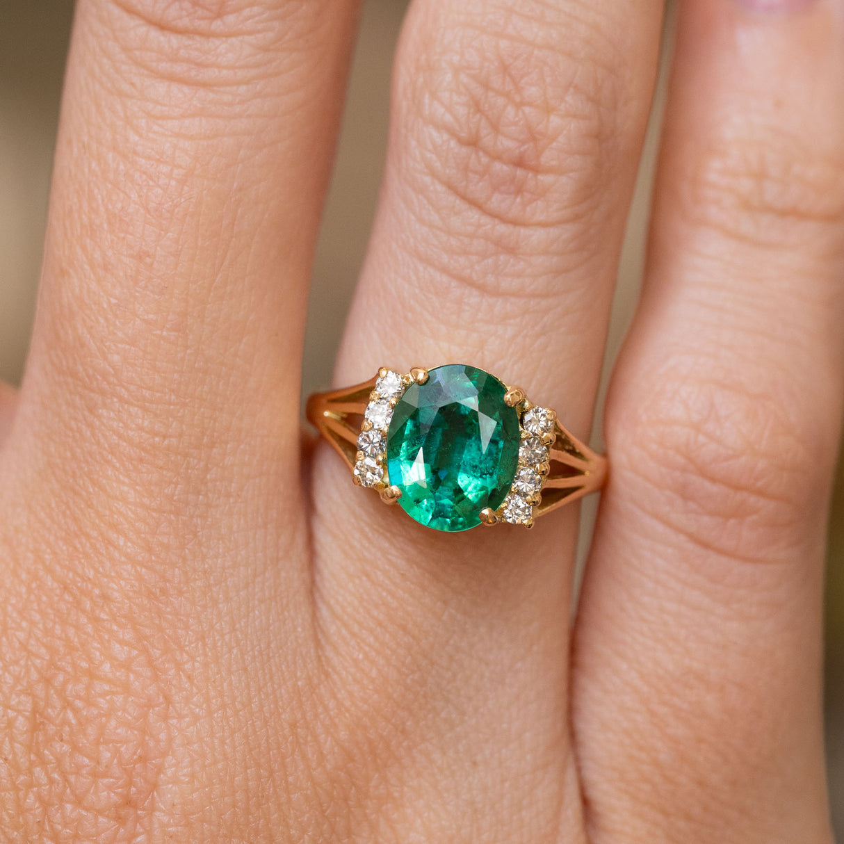 A close-up of the central emerald and flanking diamonds of a vintage 1970s engagement ring. The ring is worn on a finger and showcases the yellow gold shoulder details.