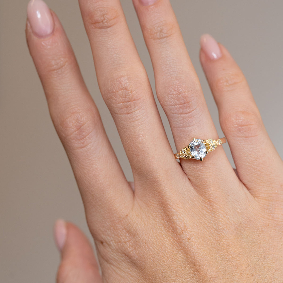 White sapphire engagement ring worn on a hand, showcasing its delicate proportions and ornate yellow gold shoulders.
