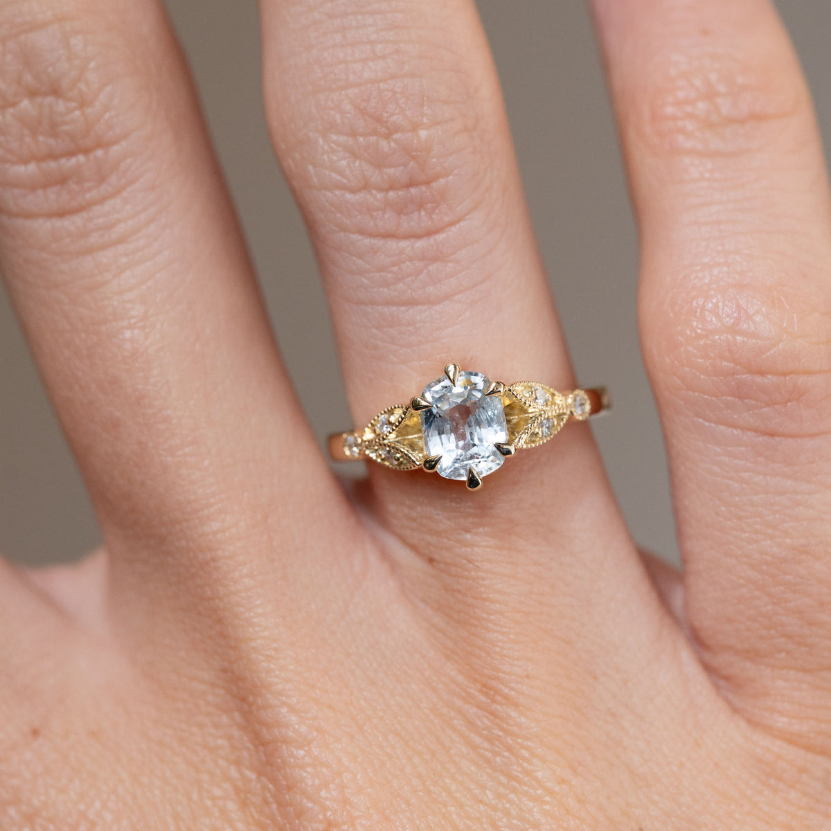 Close-up of the oval white sapphire in yellow gold, framed by decorative laurel leaf shoulders with delicate diamond accents.