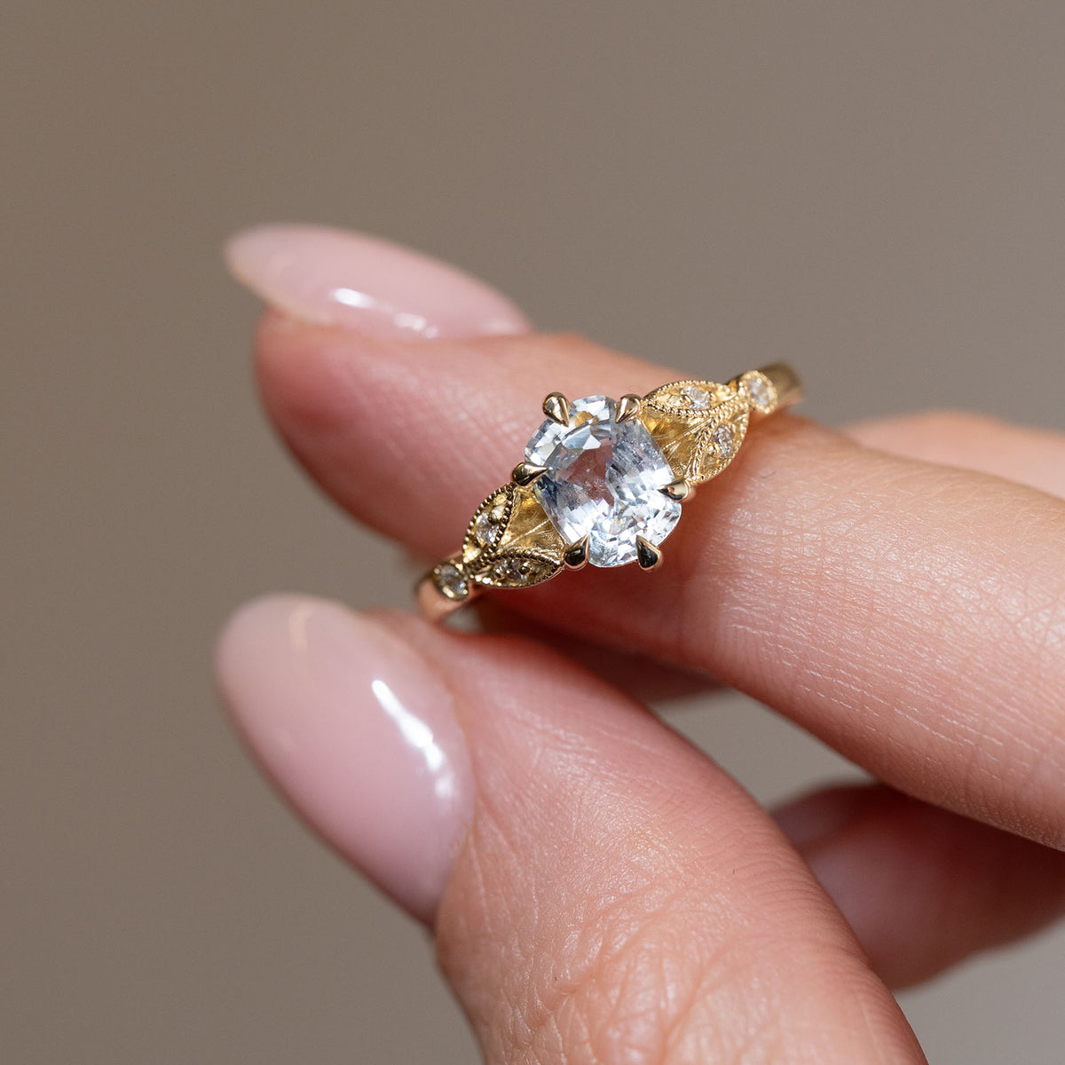 Hand-held close-up of the oval white sapphire, surrounded by delicate diamond-set leaf motifs and fine milgrain detailing.