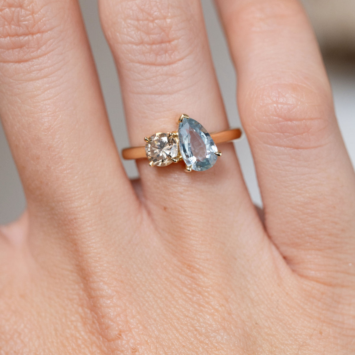 Close-up of the pear-cut blue sapphire alongside a round brown diamond, set in yellow gold and worn on a finger.