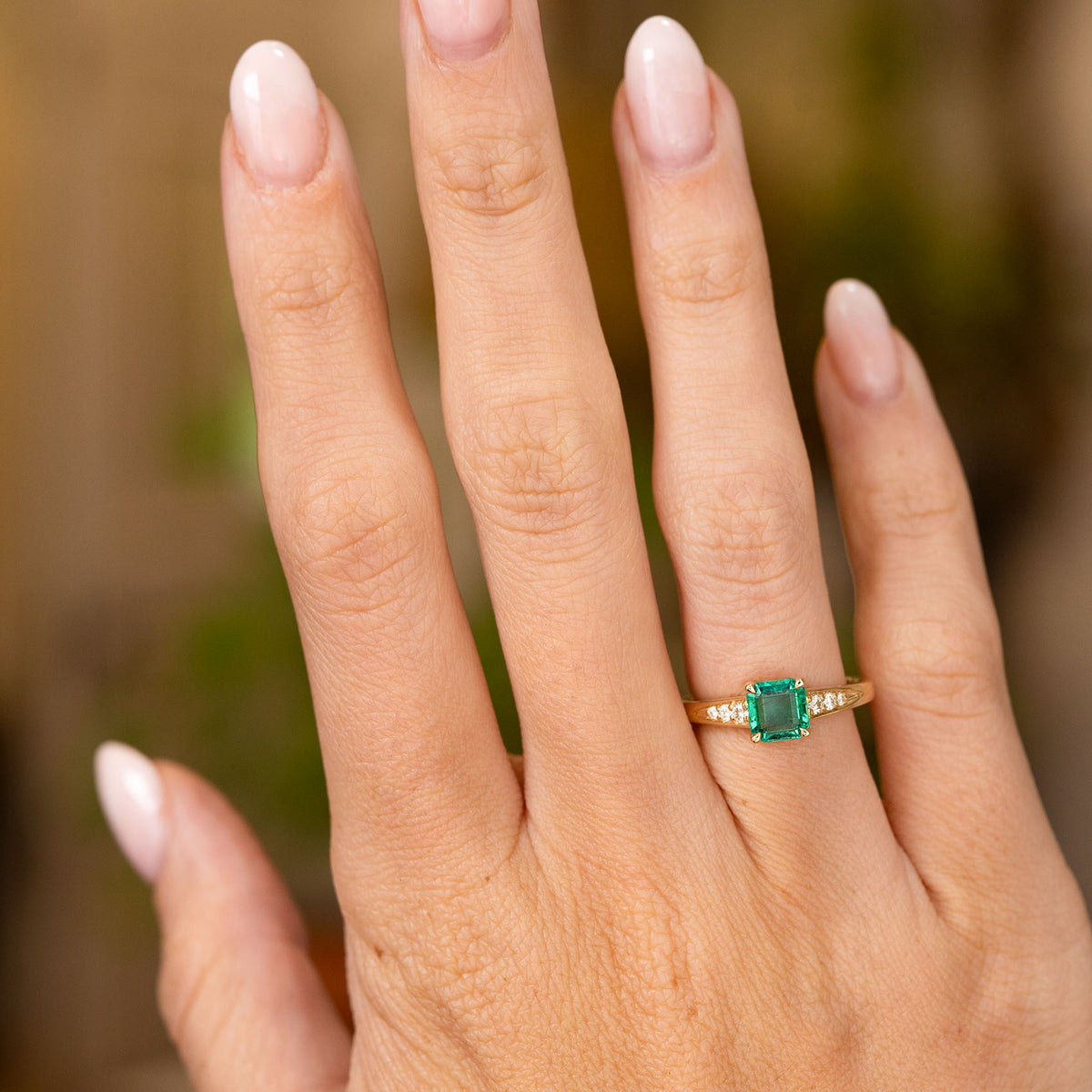 An outstretched hand wearing a yellow gold engagement ring with an emerald flanked by small diamonds shown to display the proportions