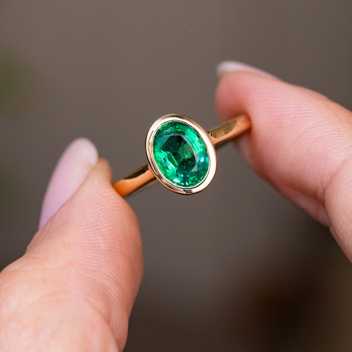 A close-up of an Antique-inspired, Emerald solitaire engagement in yellow gold being held between two fingers against a soft, blurred brown background.