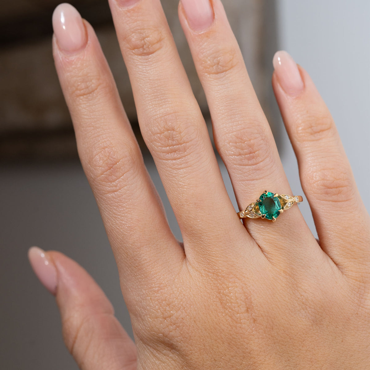Vintage-inspired emerald and diamond engagement ring worn on a hand, displaying its vibrant green centre stone and ornate laurel leaf-inspired yellow gold shoulders.