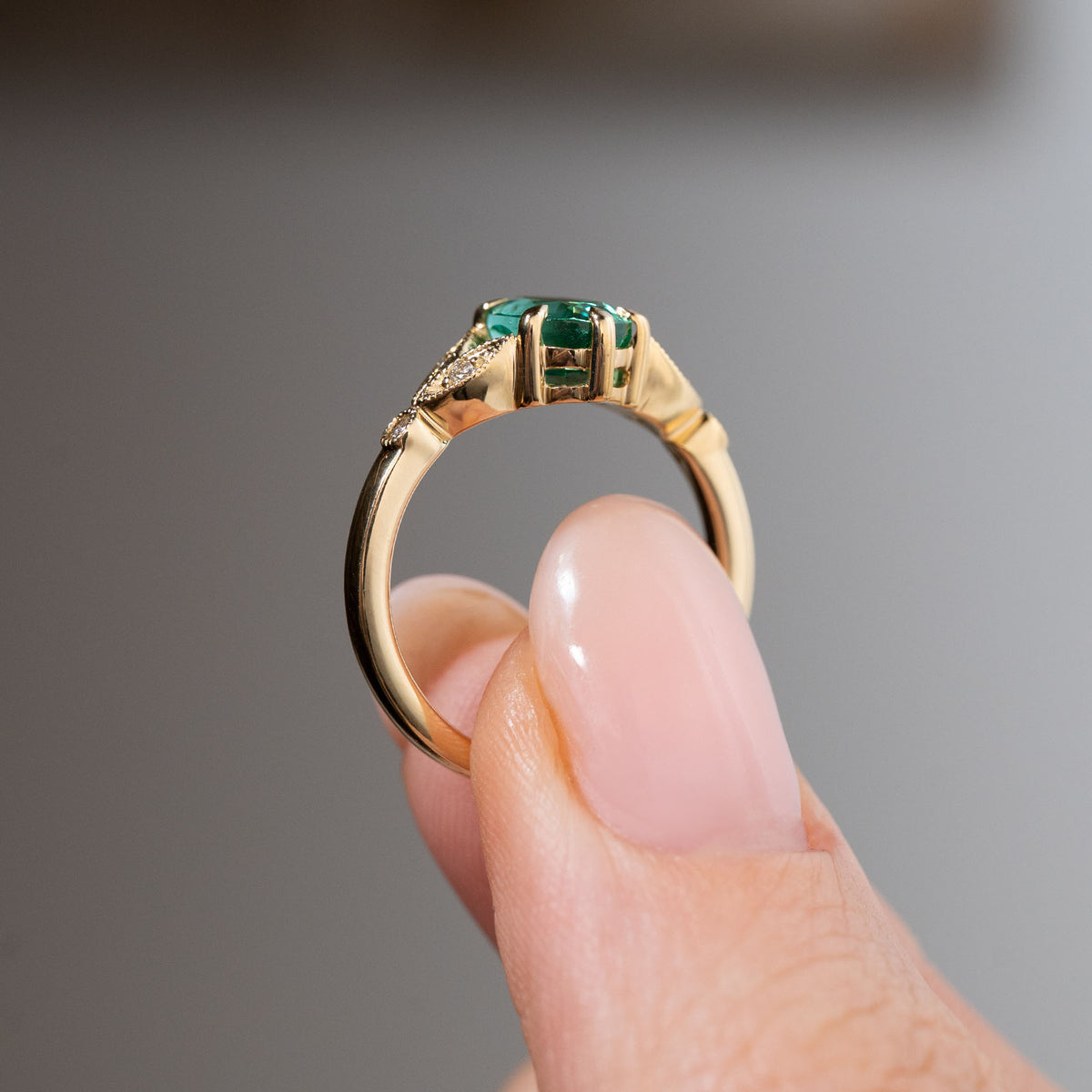 Side profile of a vintage-inspired emerald and diamond engagement ring held between fingers, showing the elevated emerald setting, sculpted yellow gold gallery and decorative trefoil shoulders.