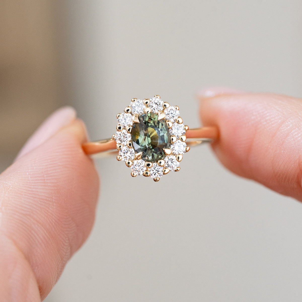 Held delicately between the fingers, the pine green sapphire contrasts beautifully with its bright round diamond halo and warm gold band.