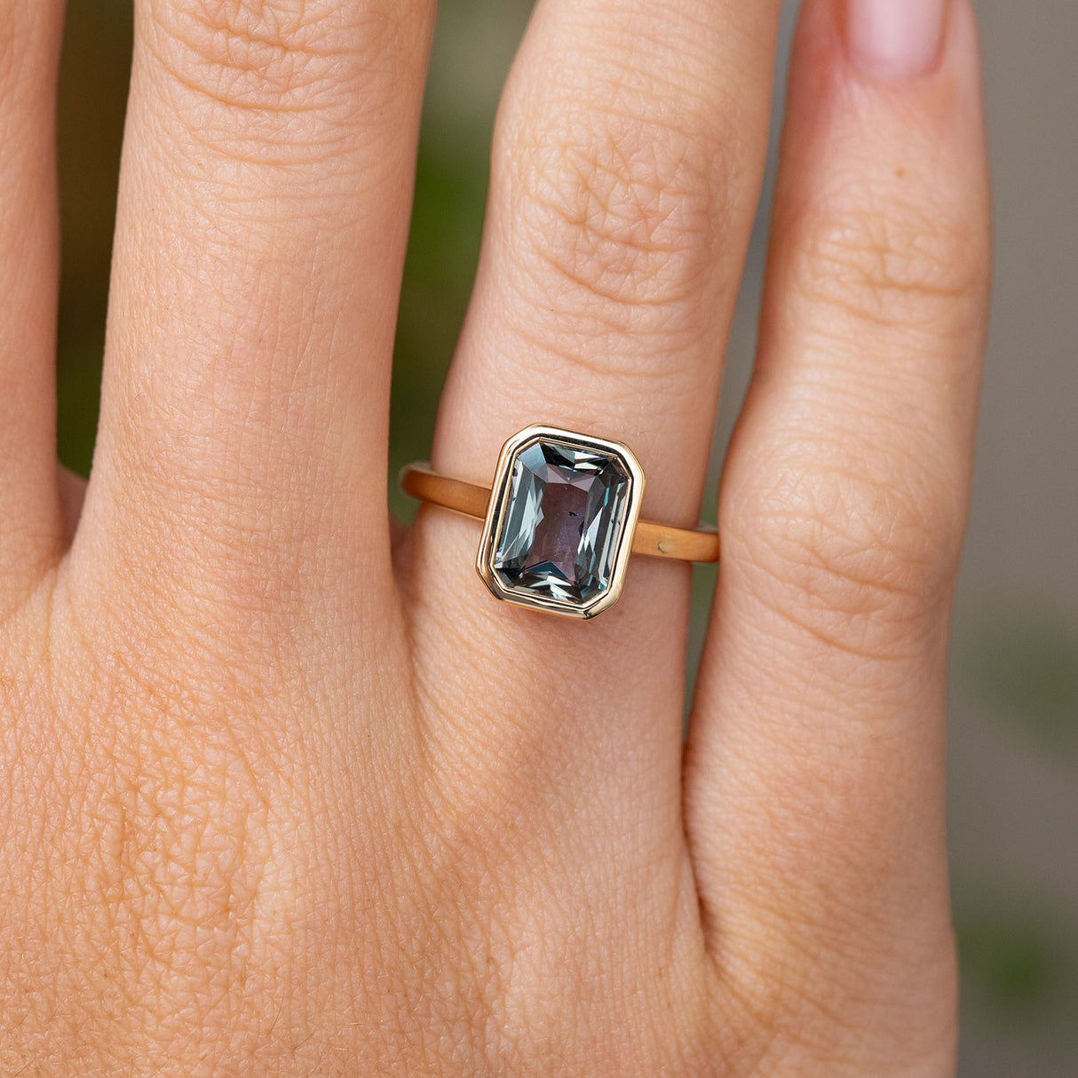 A close-up of a hand wearing a Vintage-inspired, grey-teal sapphire solitaire engagement ring. Showcasing the yellow gold band and rub-over bezel setting of the sapphire.