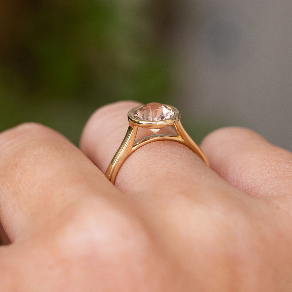 A close-up of a yellow gold solitaire engagement ring worn on a person's finger against a blurred green and gray background. Displaying the setting and profile of the pale pink round sapphire.