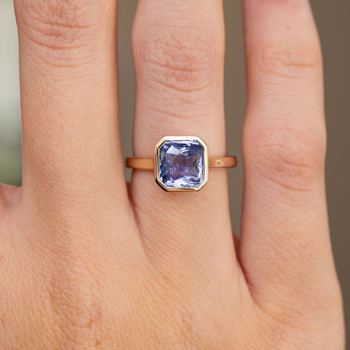 Close-up on-hand view of the emerald-cut lavender sapphire within a sleek geometric gold surround.