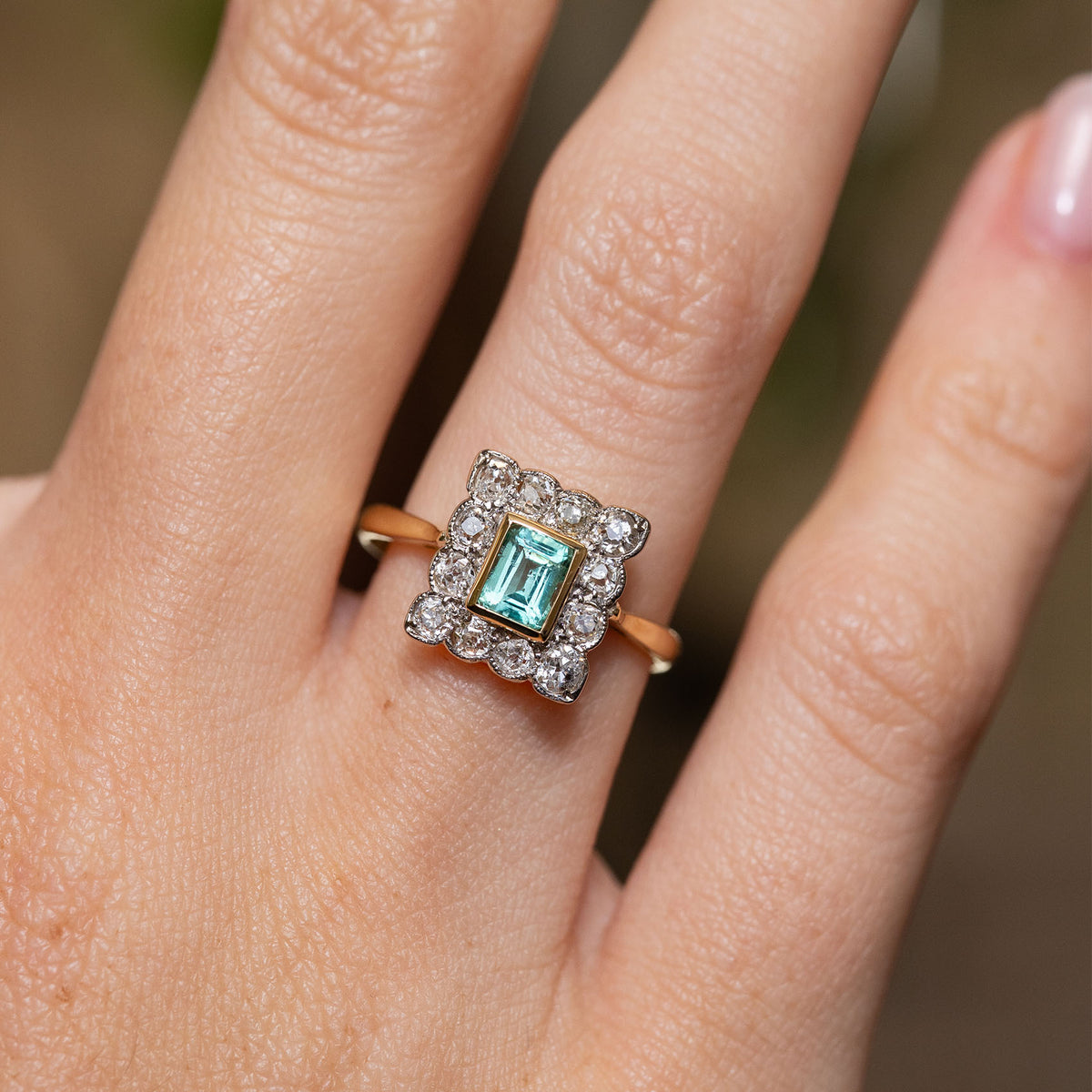 Close-up of an antique emerald and diamond engagement ring worn on the hand, showing its yellow gold bezel and scalloped platinum cluster.