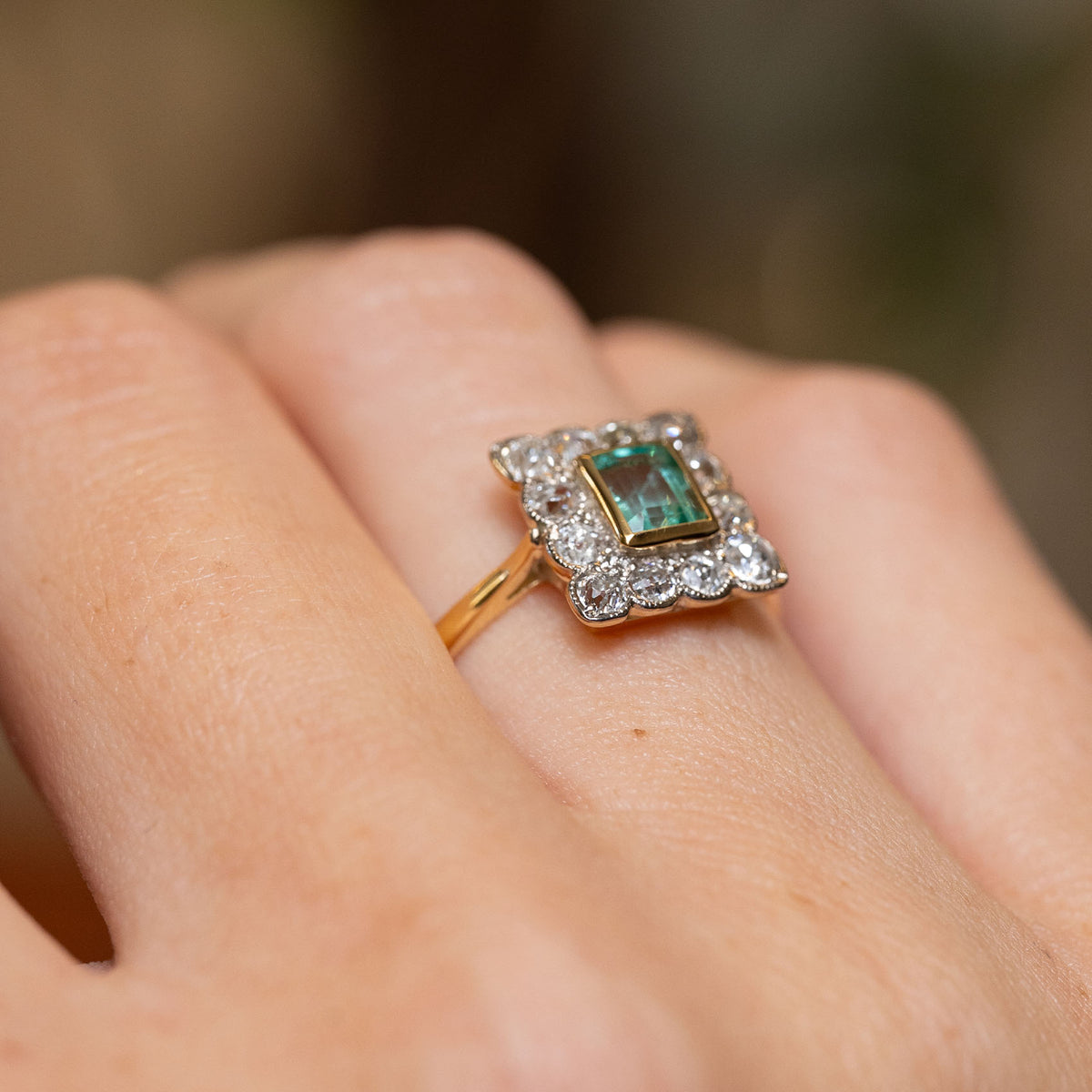 Angled hand view of a mint green step-cut emerald ring with an ornate old-mine cut diamond border in platinum.