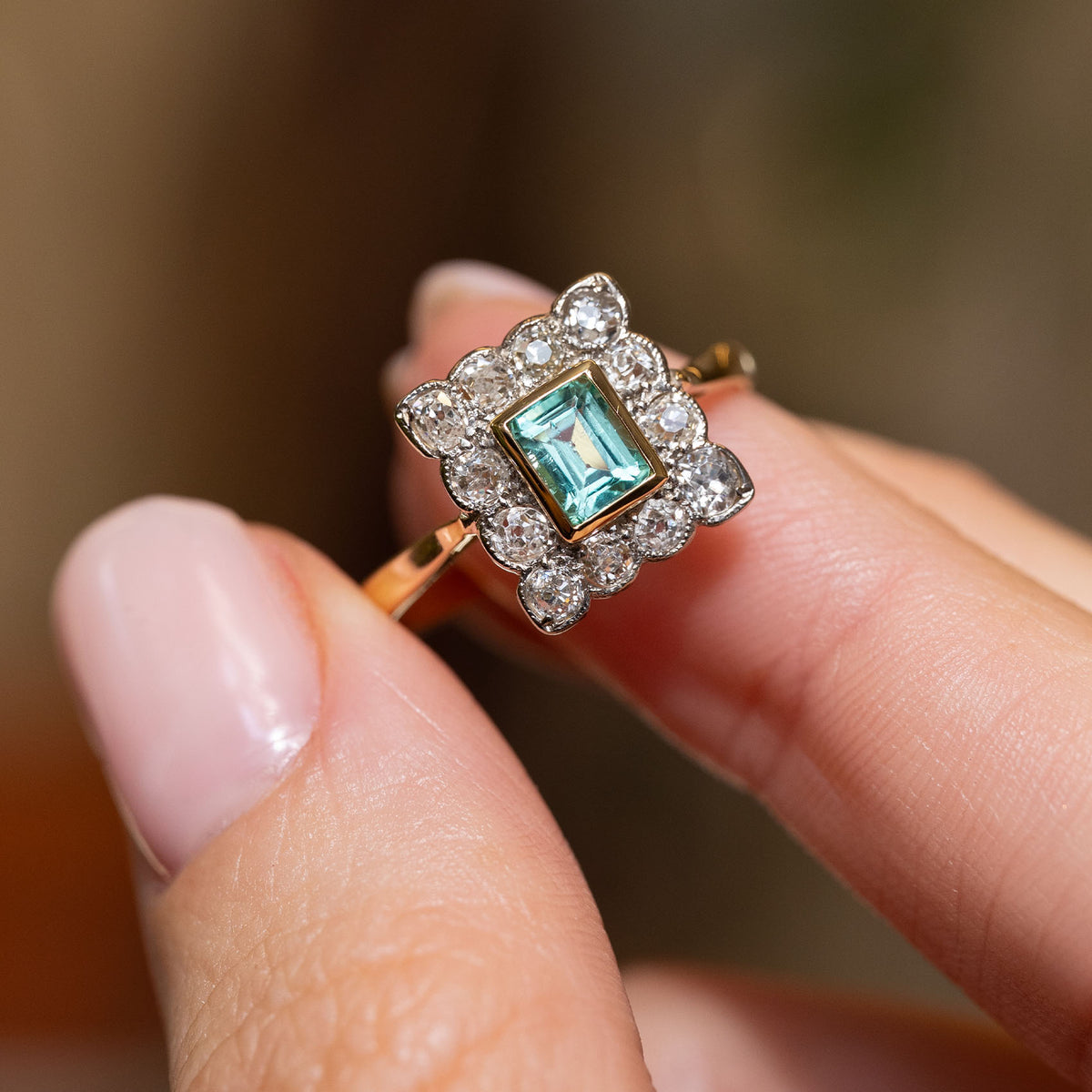 Antique Edwardian emerald and diamond ring held between the fingers, showcasing its geometric cluster and contrasting yellow gold and platinum setting.