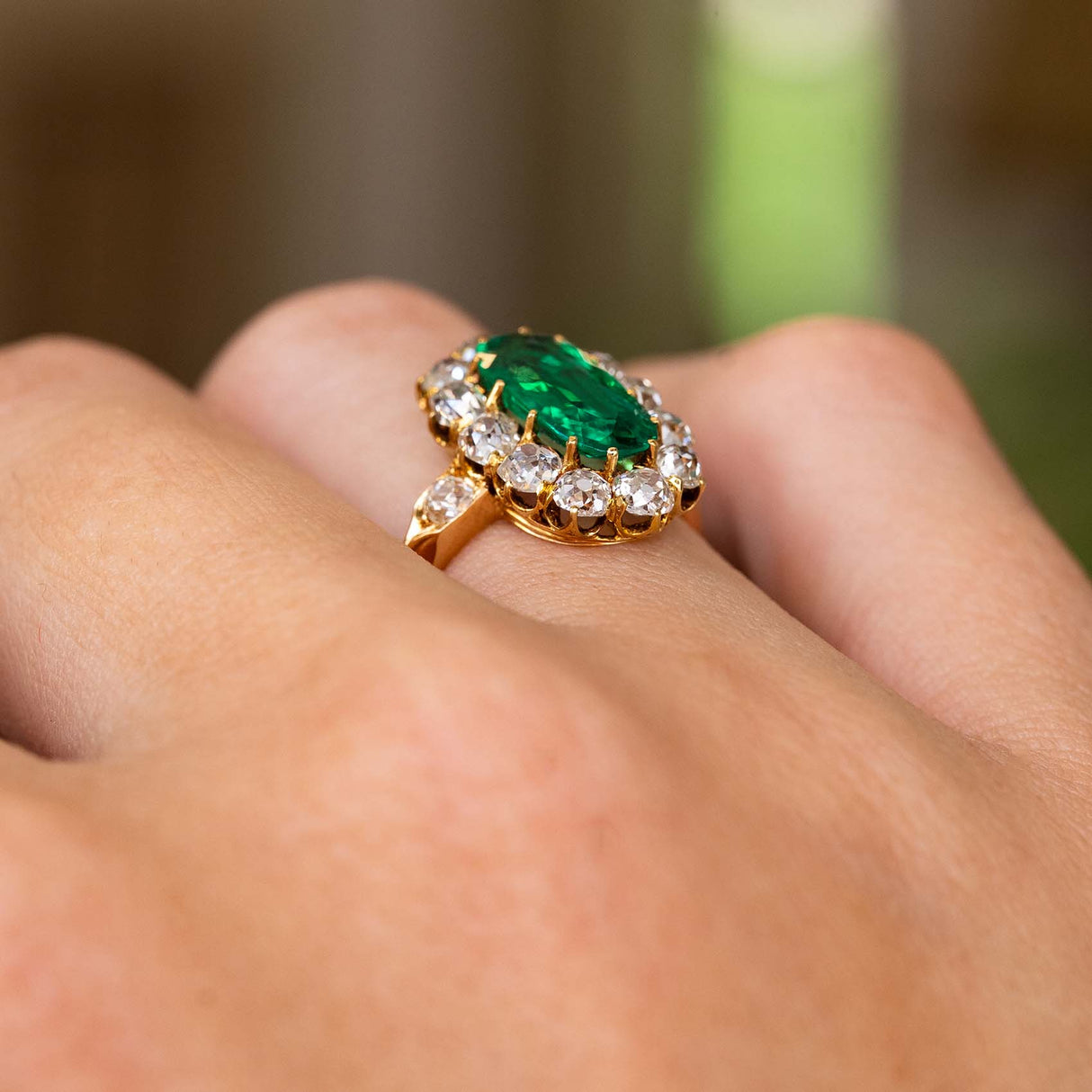 A close up of the side profile of an Antique, Belle Époque Colombian emerald and diamond cluster engagement ring. Showing the profile of the ring once worn, the yellow gold band and shoulder details.