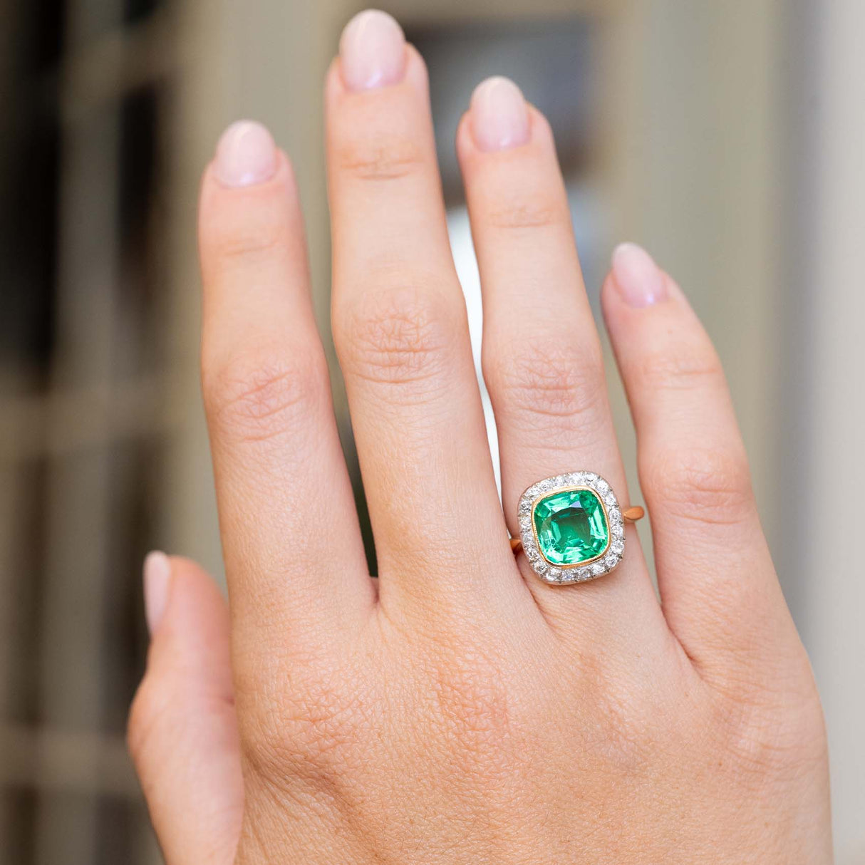 An image of a hand outstretched wearing an antique Belle Époque era yellow gold engagement ring featuring an emerald and diamonds worn on the finger against a blurred background.