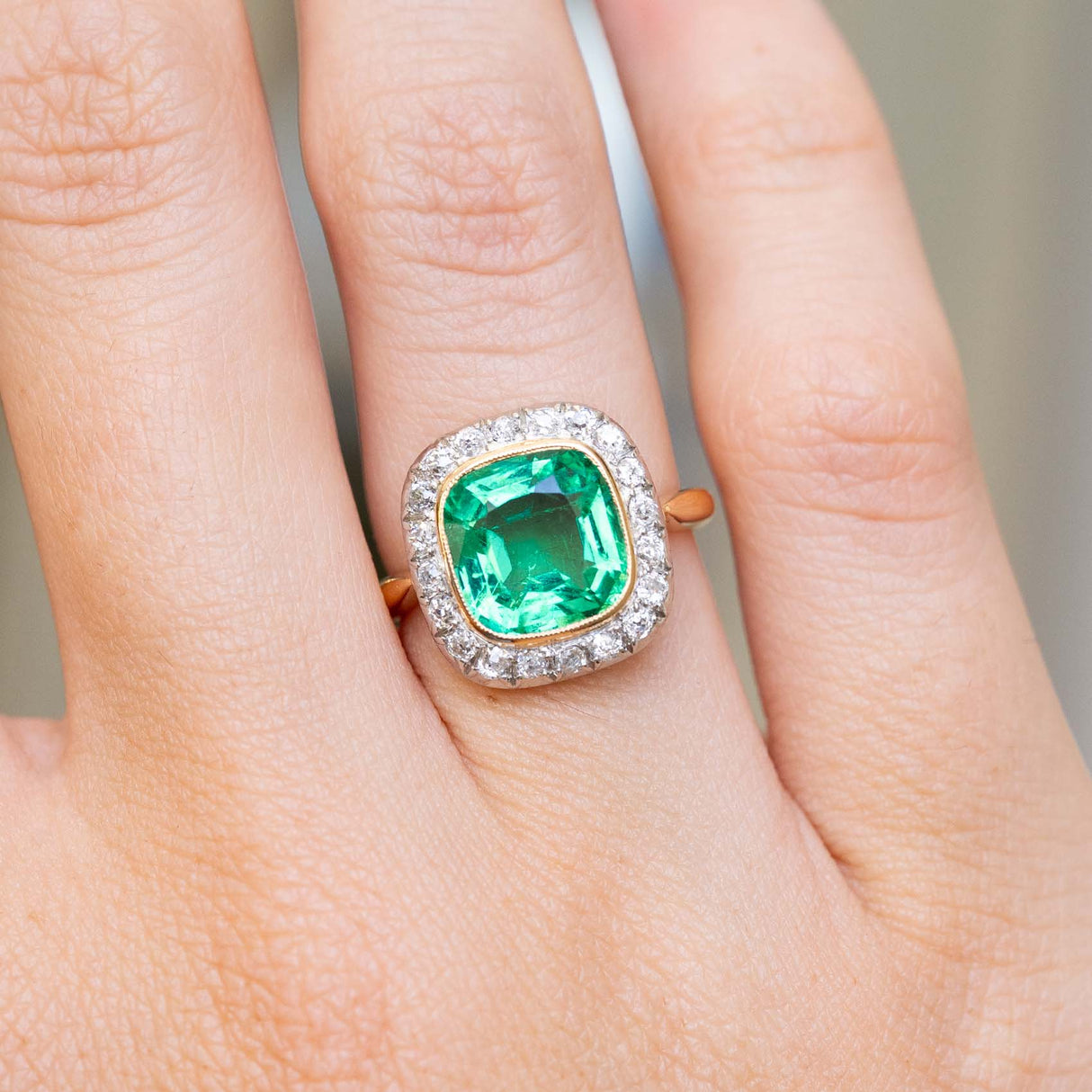 A close-up picture of an antique Belle Époque era emerald and diamond coronet cluster yellow gold engagement ring worn on the finger and held up to show details.