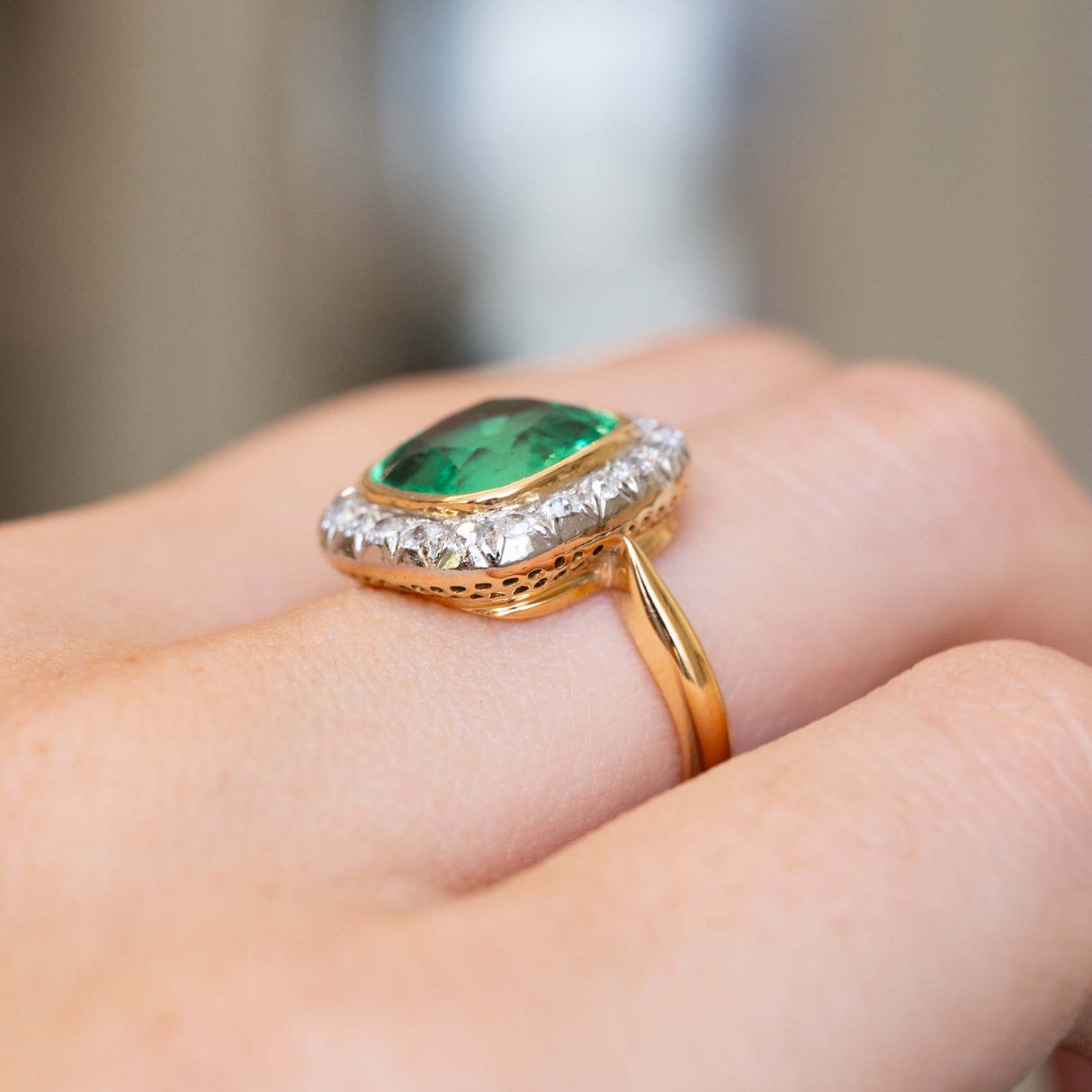 A zoomed in image of an antique Belle Époque era emerald and diamond coronet cluster yellow gold engagement ring worn on the finger and shown at an angle to display how the ring sits on the finger when worn.