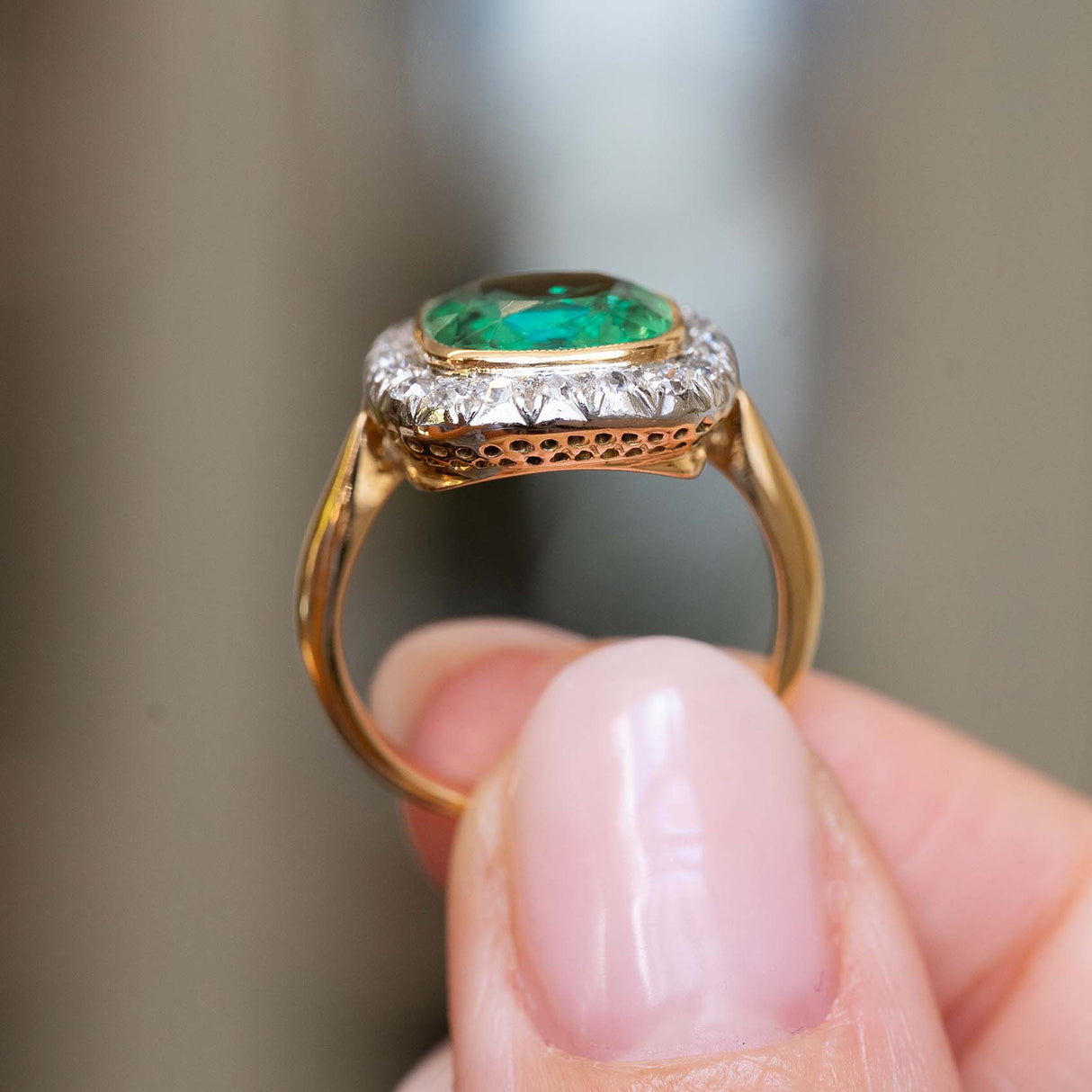 An antique Belle Époque cushion-cut emerald and diamond cluster engagement ring in yellow gold held up between the fingers to show the intricately pierced gallery and setting style of the diamonds.