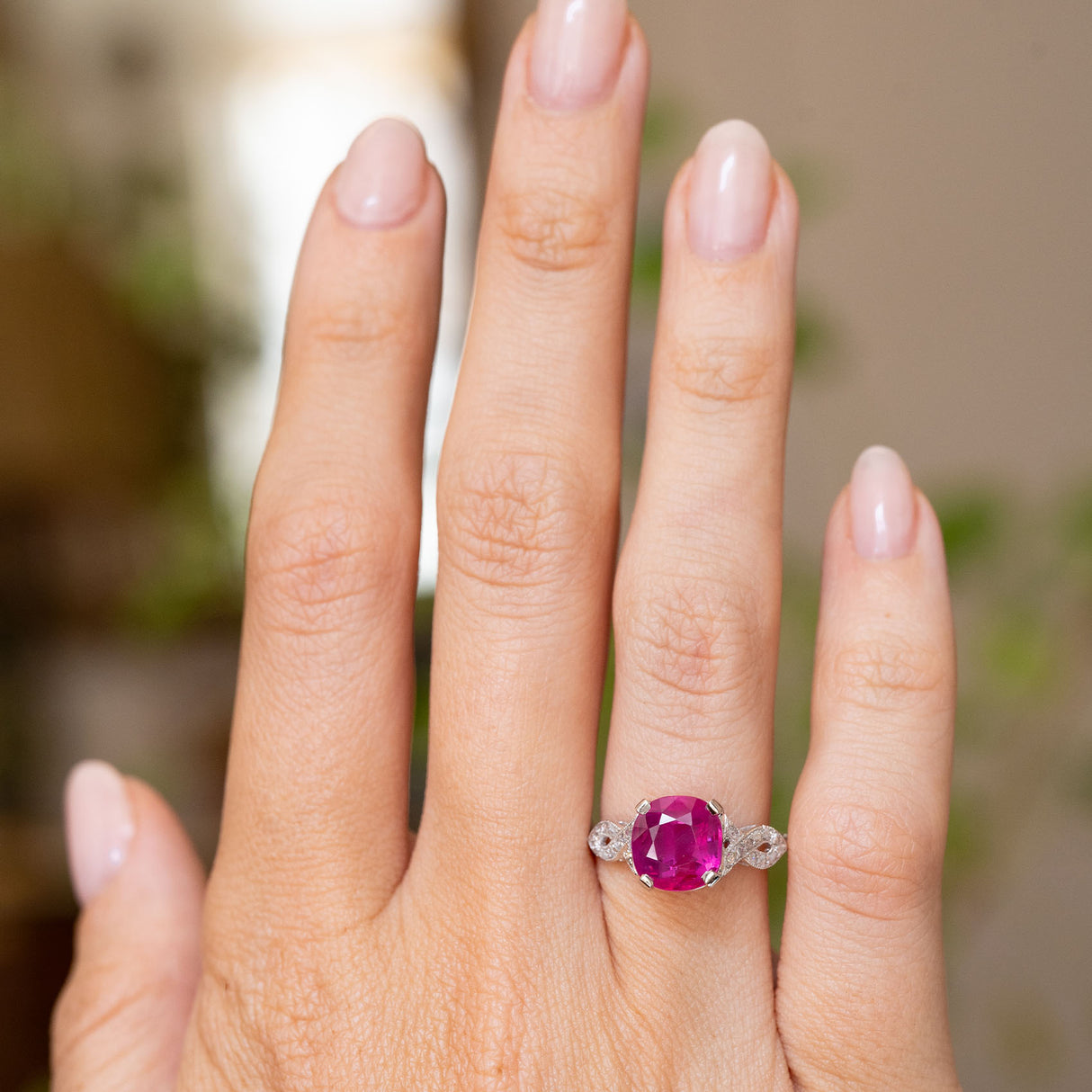 A close-up of a hand wearing a white gold Antique engagement ring from the Art Deco era, featuring a central cushion-cut pink ruby flanked by smaller diamonds on either side.