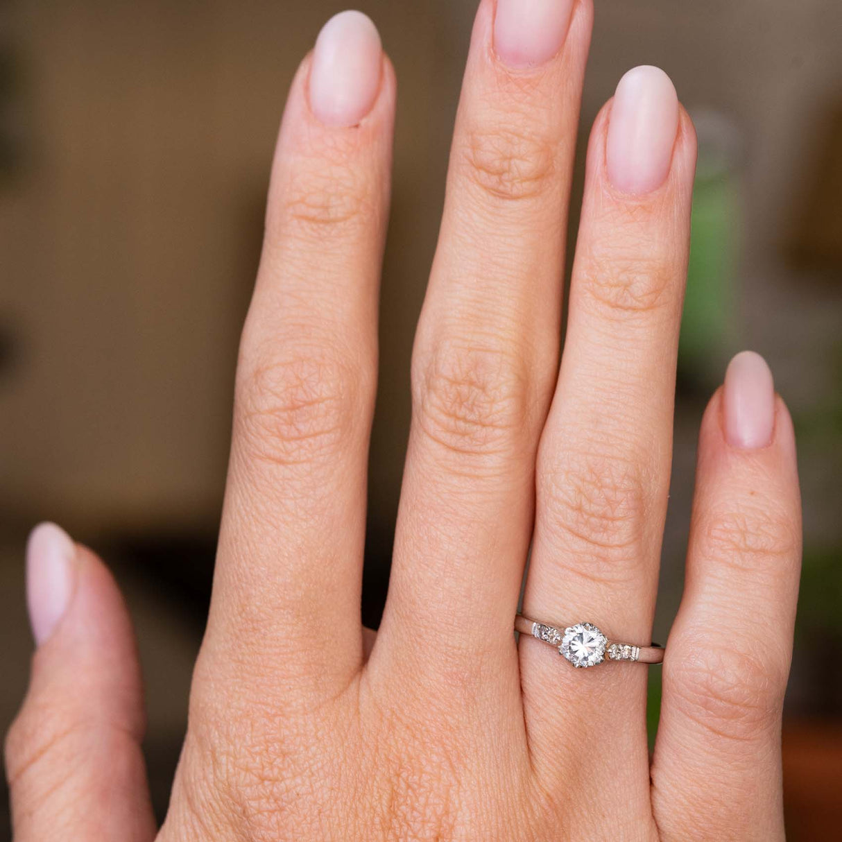 A close-up of a hand wearing a platinum diamond engagement ring with a round-cut central diamond and smaller flanking diamonds. The ring is Antique from the 1930s Art Deco era.