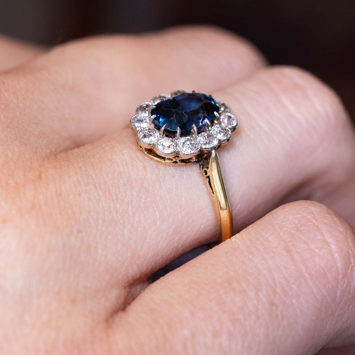 A close-up image of a yellow gold Antique, Edwardian blue spinel and diamond cluster engagement ring in yellow gold worn on a finger and seen from the side to show the shoulders and setting style of the ring.