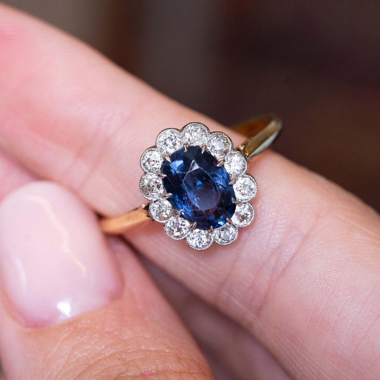 A picture of an Antique, Edwardian dark blue oval-cut spinel and diamond coronet cluster engagement ring on a yellow gold band balanced between the finger and the thumb, seen against a blurred background.