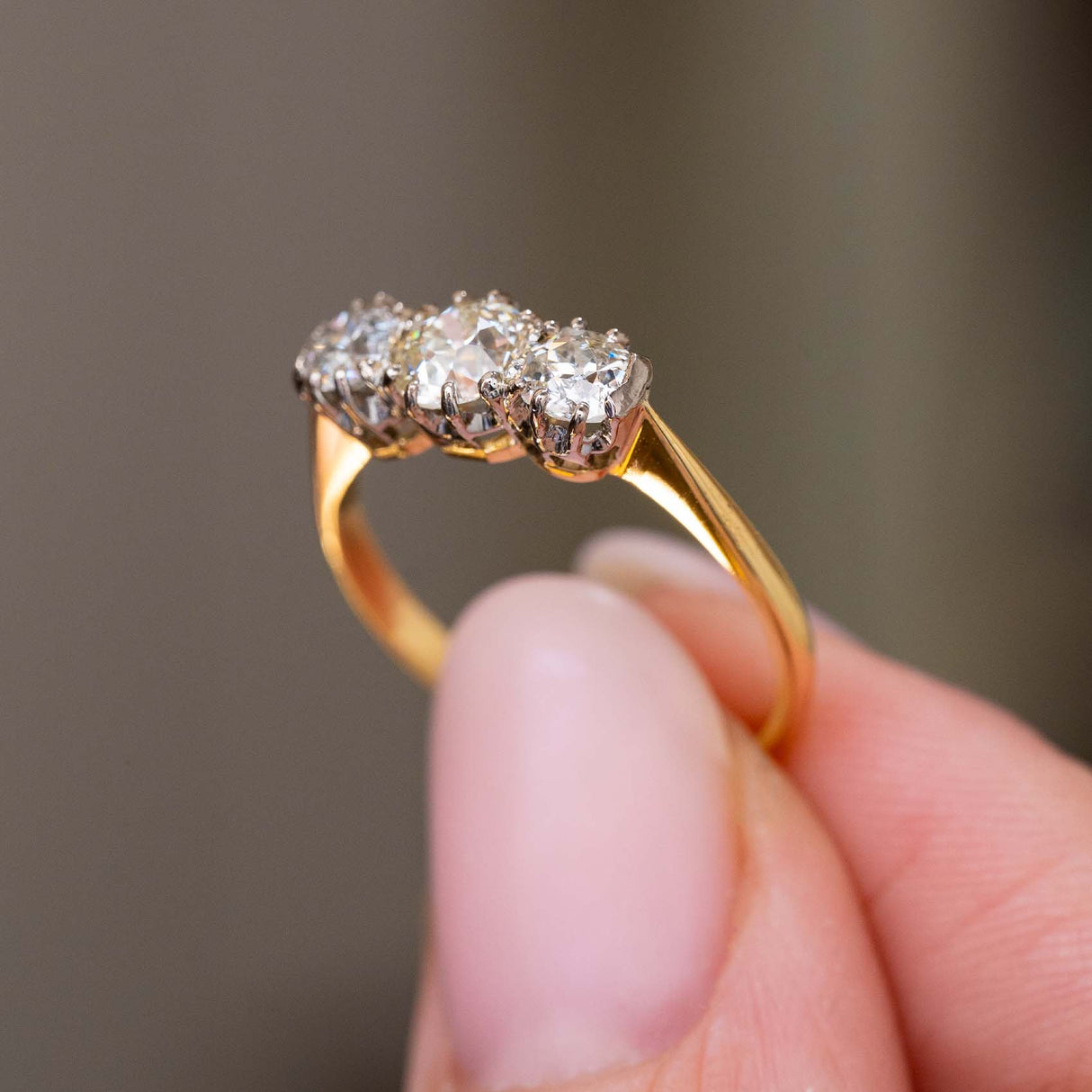 A close-up of a yellow gold Antique, Edwardian diamond three-stone engagement ring held between fingers to show the gallery of the ring against a soft, blurred grey background.