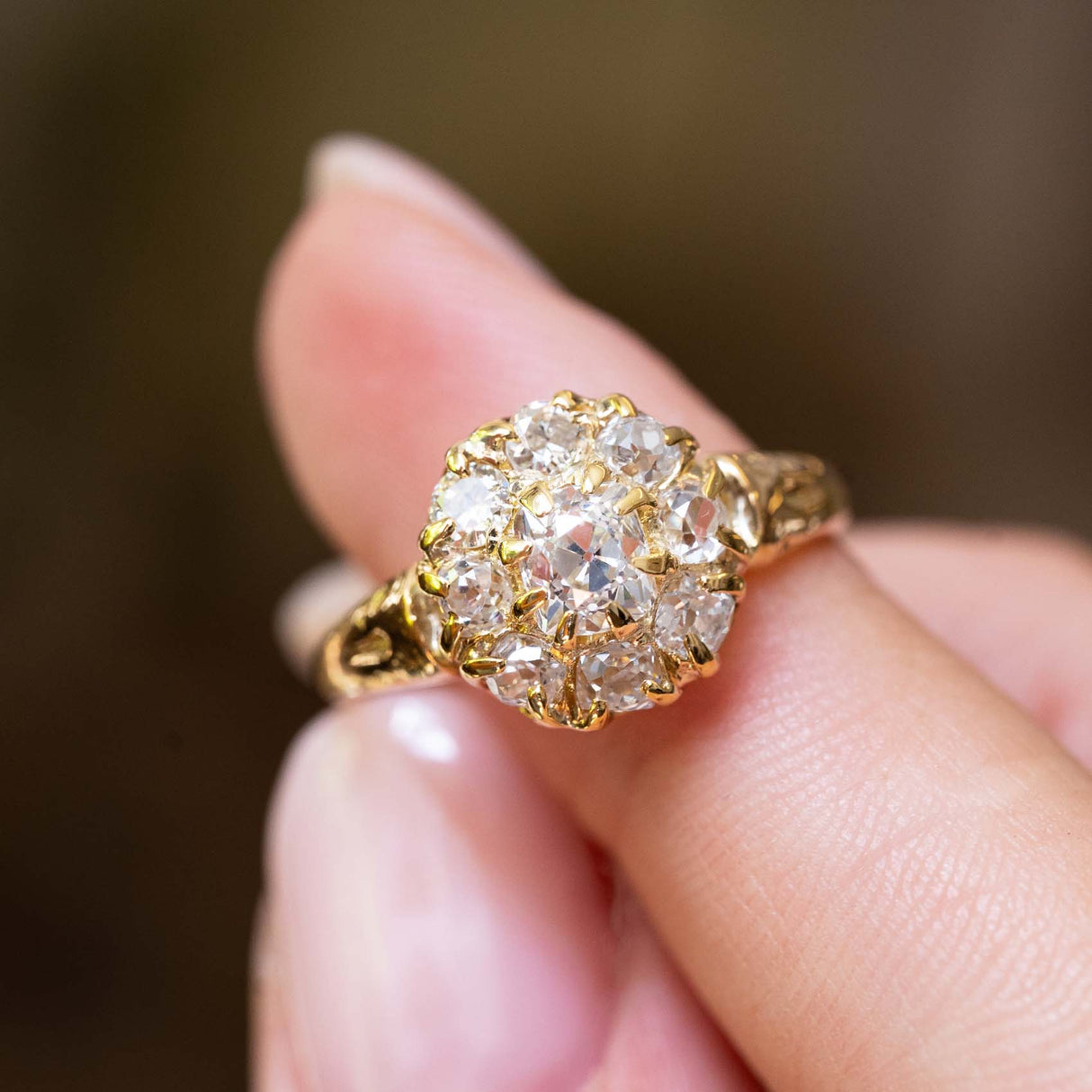 A close-up of an antique Victorian era yellow gold coronet cluster ring balanced on a person's finger against a softly blurred brown background.