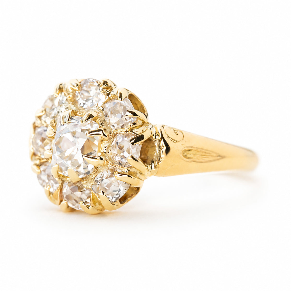 An antique yellow gold cluster engagement ring with old cut diamonds and engraved shoulders shown from the side against a plain white background.