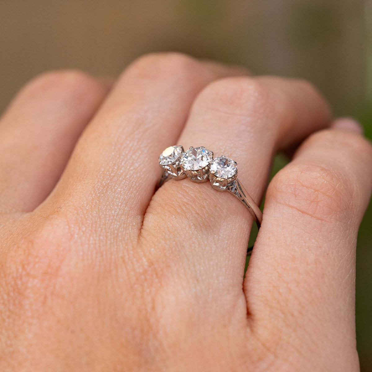 A close-up of a hand wearing a platinum Antique, Edwardian diamond three-stone engagement ring with the fingers resting against a soft, blurred green background.