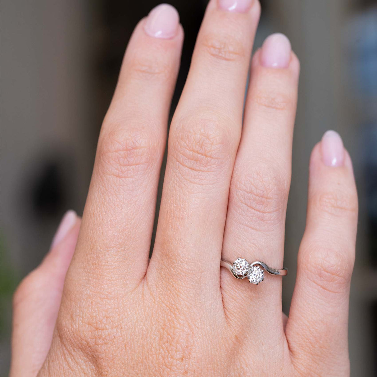 A photo of a hand wearing an Antique, Edwardian toi et moi style two stone diamond engagement ring in platinum against a softly blurred background, showing the proportions of the ring when worn.