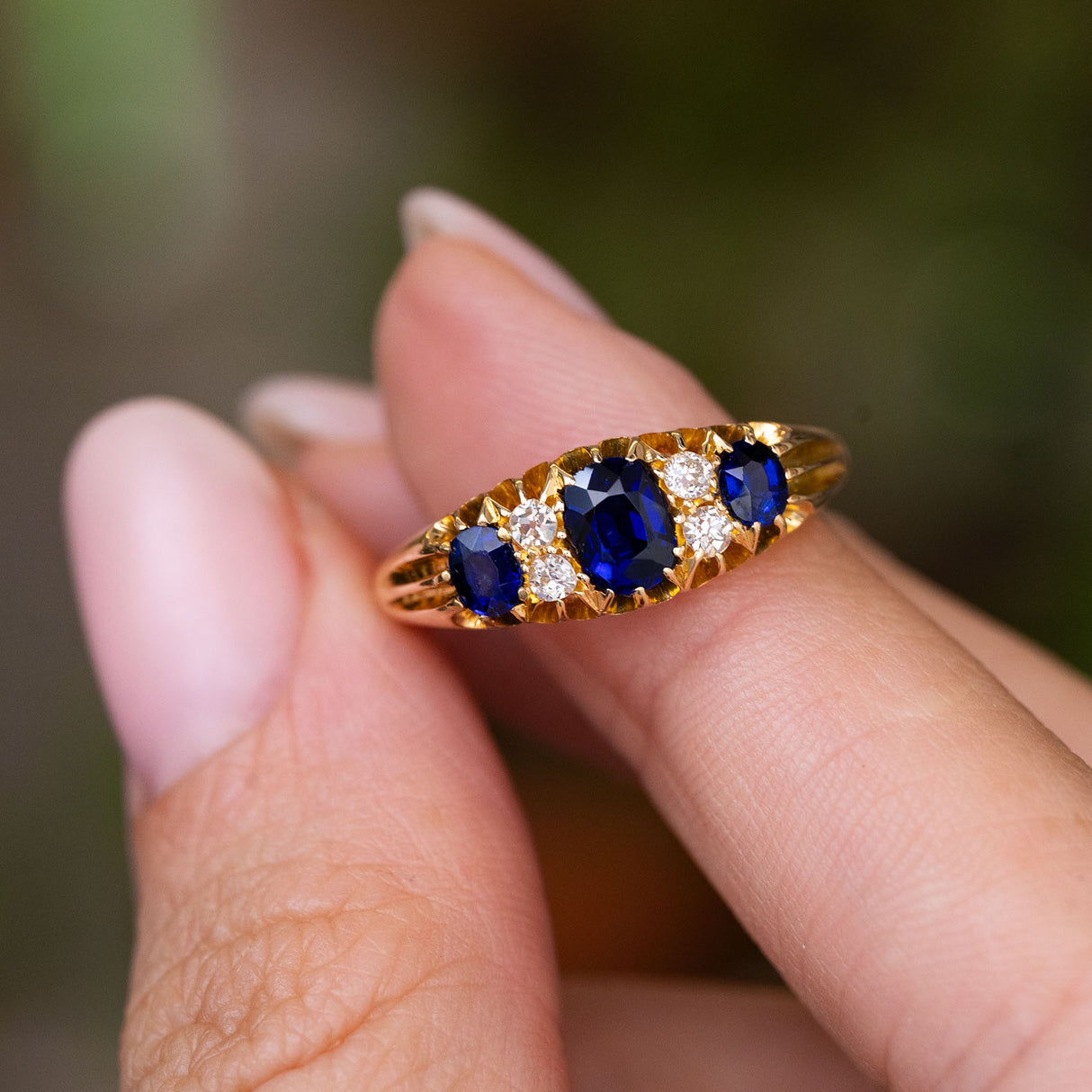 A yellow gold antique Edwardian five-stone engagement ring set with three cushion-cut blue sapphires and smaller accent diamonds, held between fingers against a blurred green background.