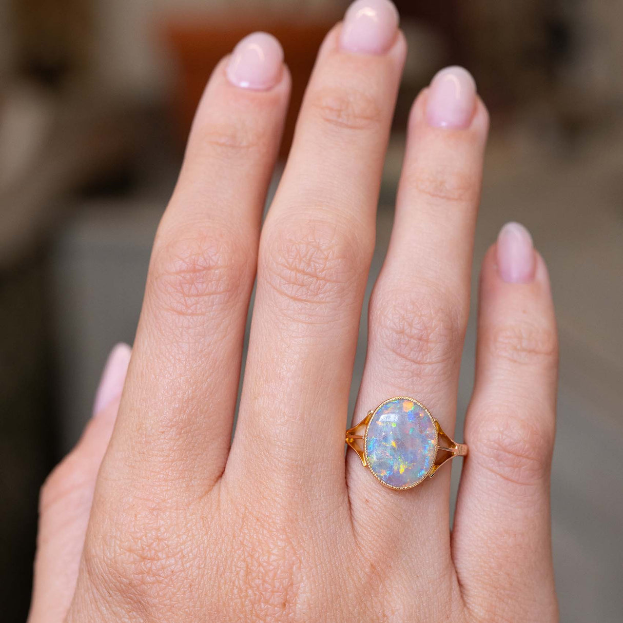A close-up picture of an outstretched hand wearing a gold ring with an oval opal gemstone in yellow gold mount displayed at a distance to showcase the scale of the ring when worn.